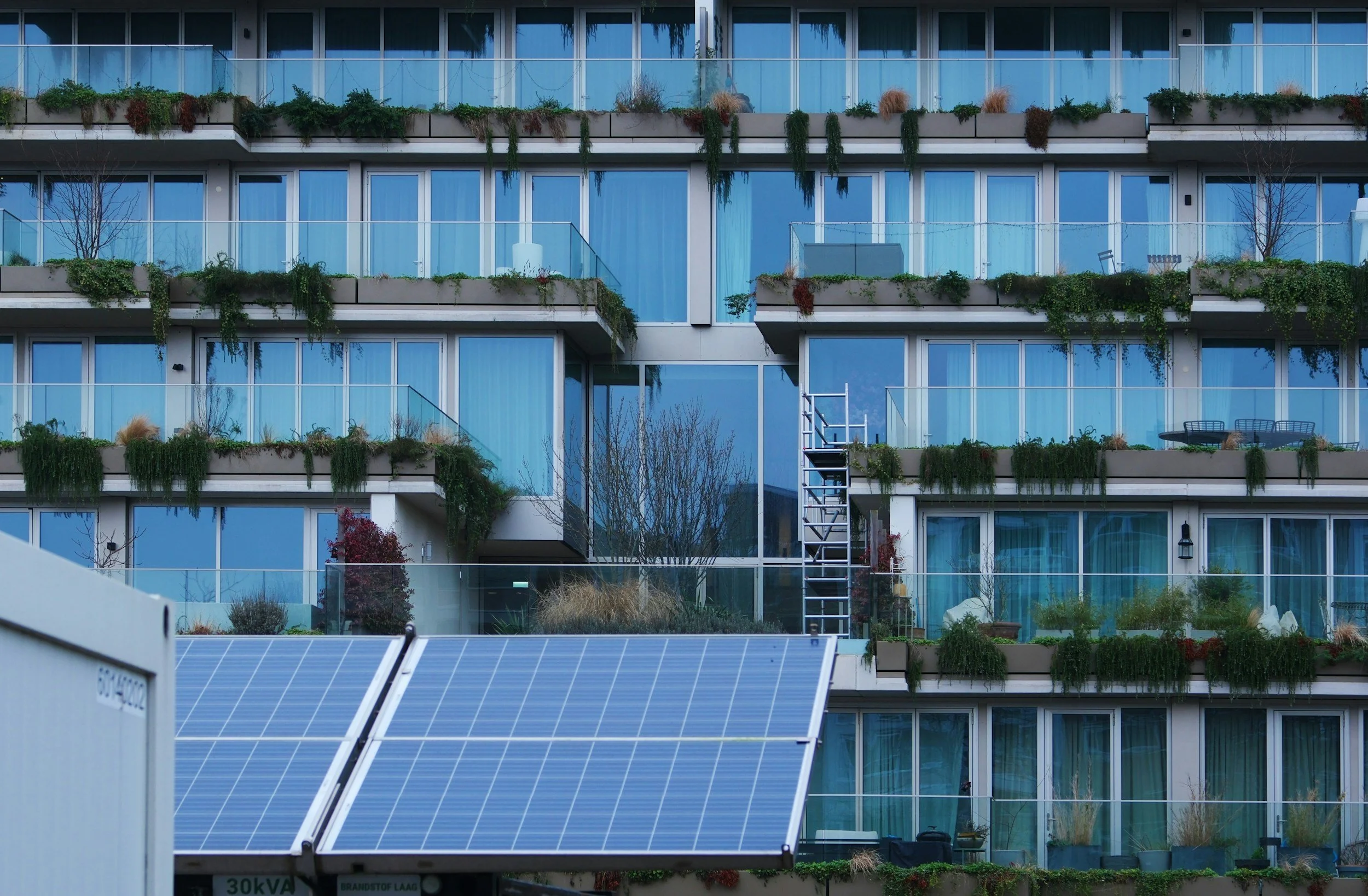 A modern multi-story building with extensive glass windows and balconies, featuring rooftop and balcony gardens with various plants. Solar panels are visible in the foreground.