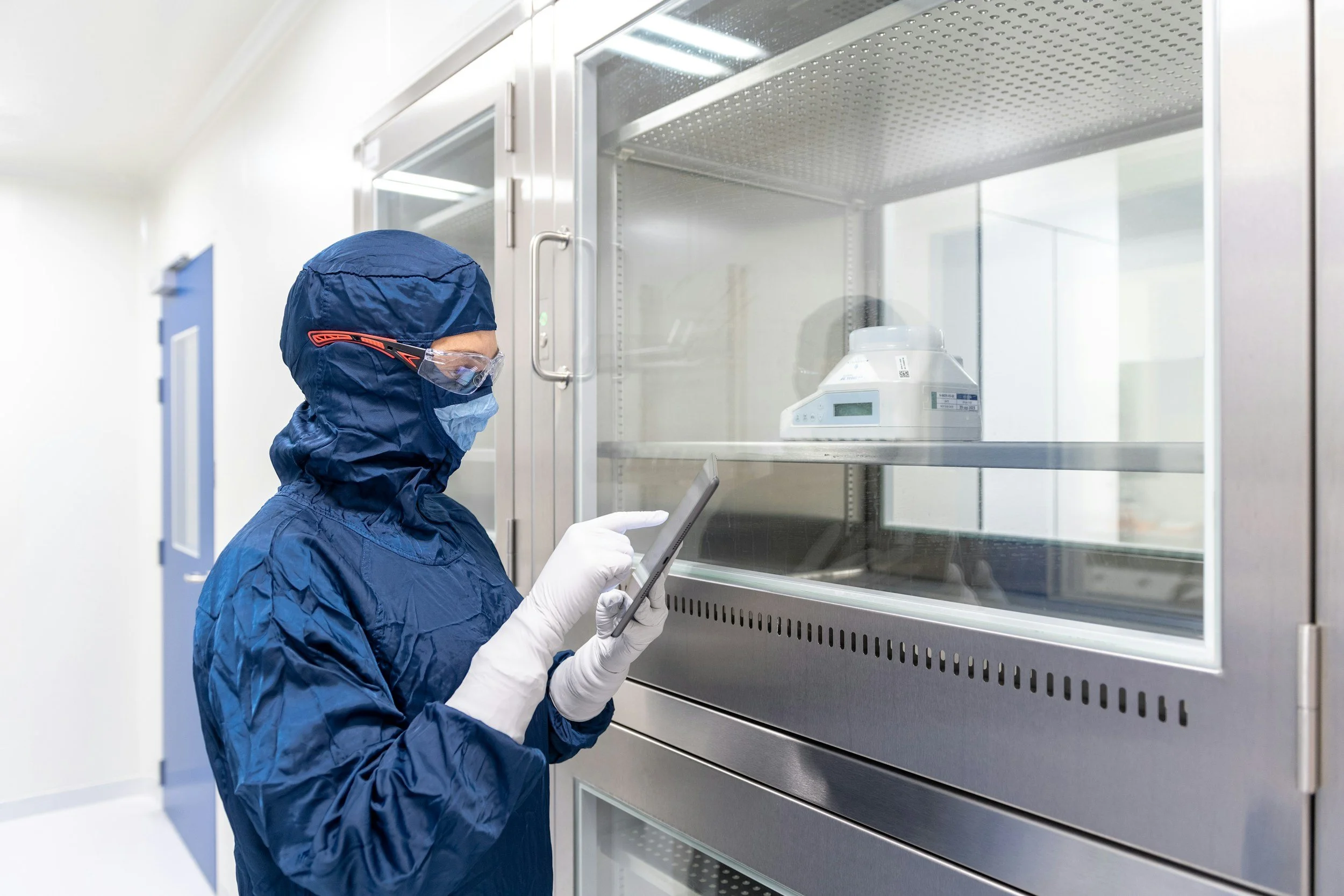 A scientist in protective gear, including a blue hooded suit, mask, goggles, and gloves, using a tablet in a laboratory with a sealed cabinet containing scientific equipment.