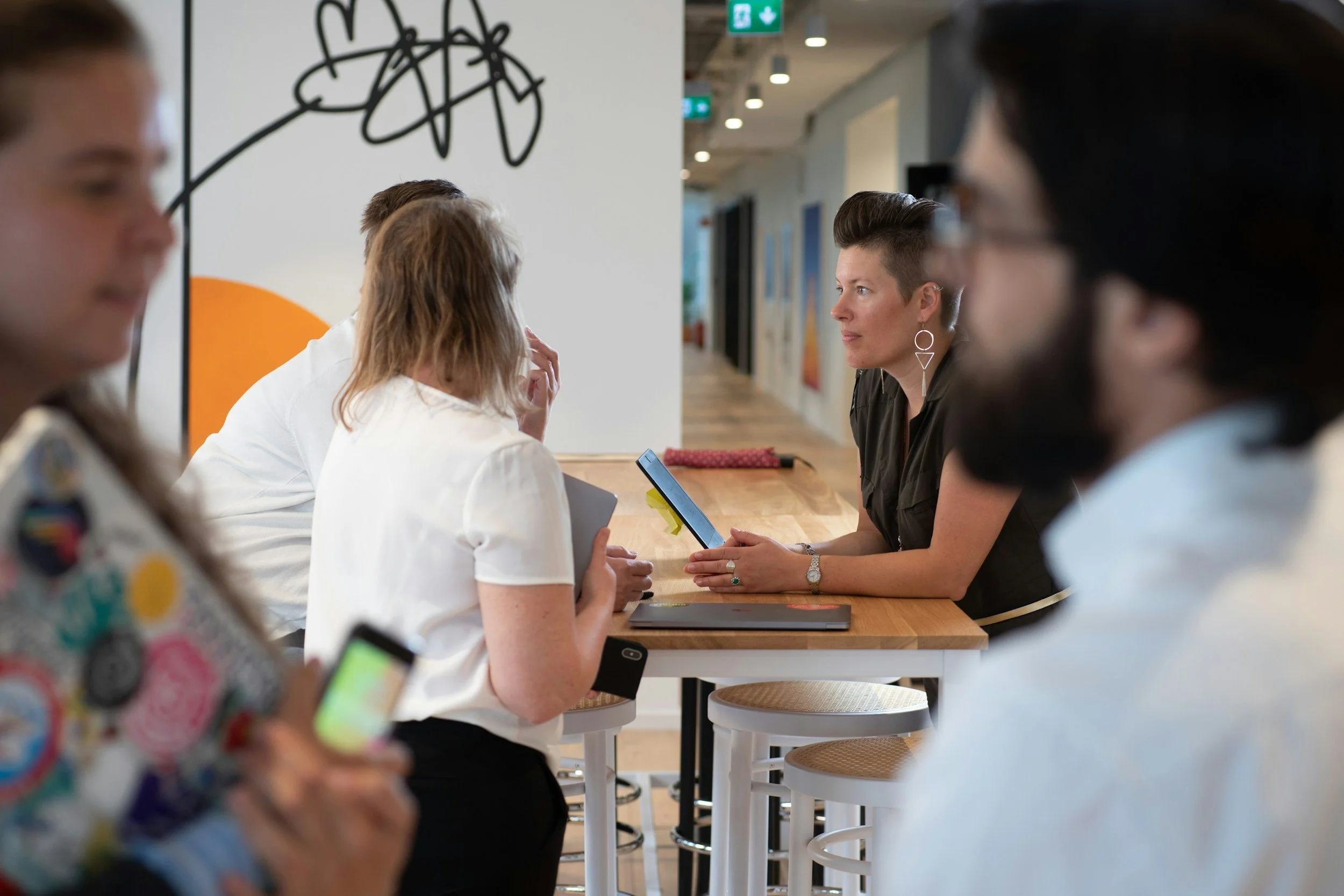 A group of diverse people sitting at a high table in what appears to be an art gallery or modern office space, engaged in conversation, with some holding electronic devices.