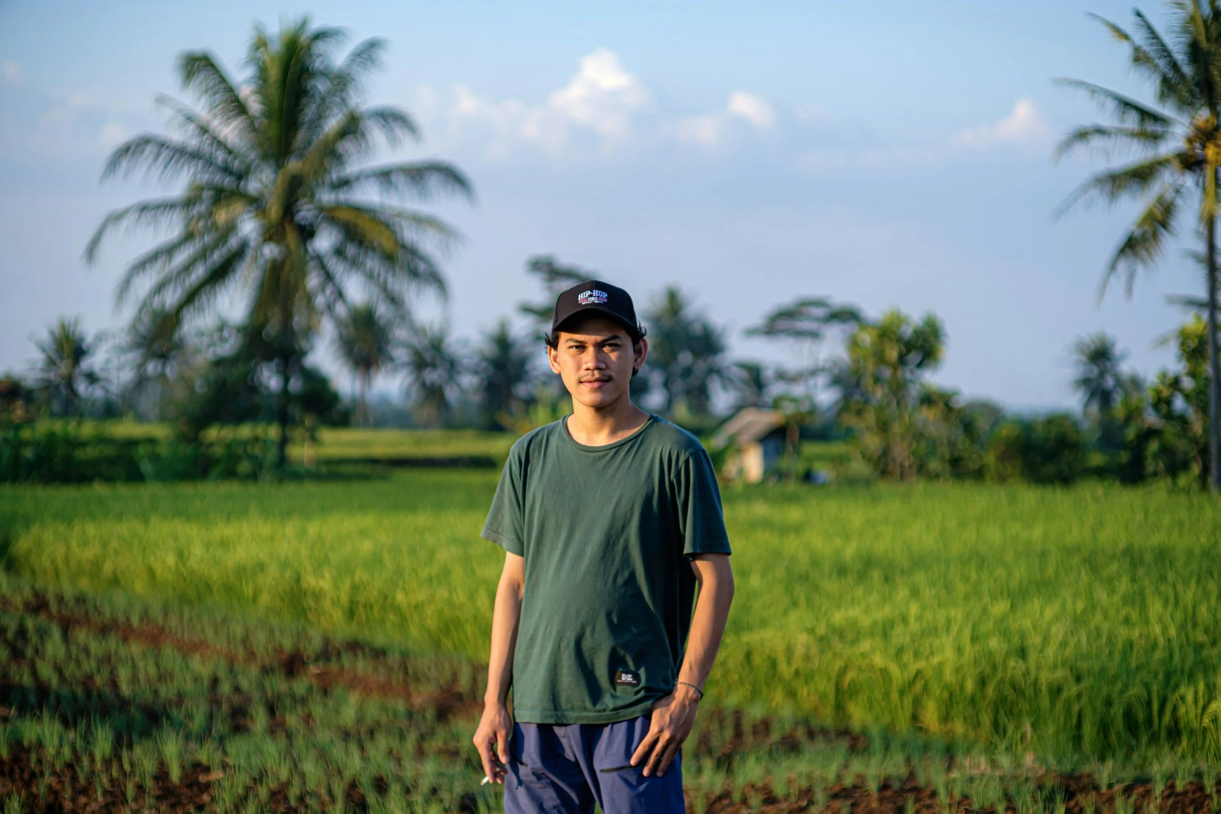 A young man in a green t-shirt and a black cap stands in a lush green rice field with tall palm trees and a small structure in the background, under a partly cloudy sky.