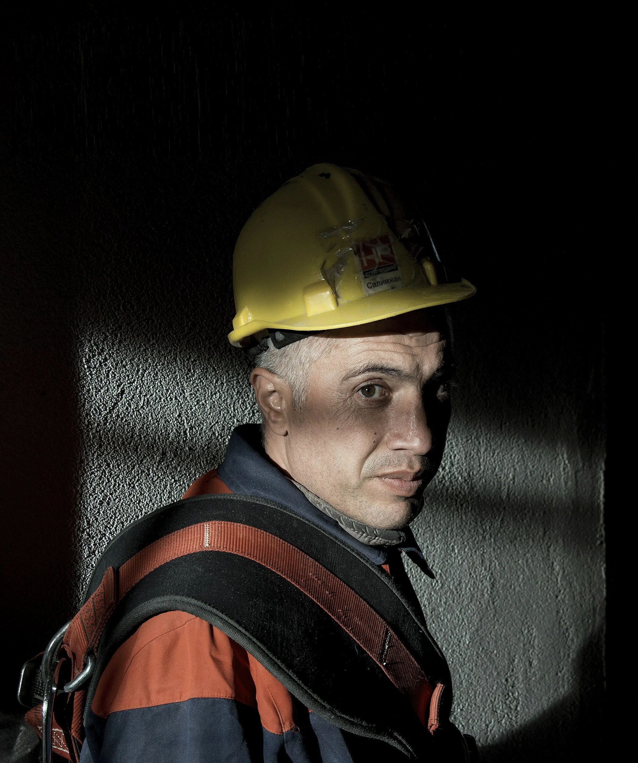 A male construction worker with a yellow safety helmet and orange and black work gear, standing against a dark wall with lighting casting shadows on his face.