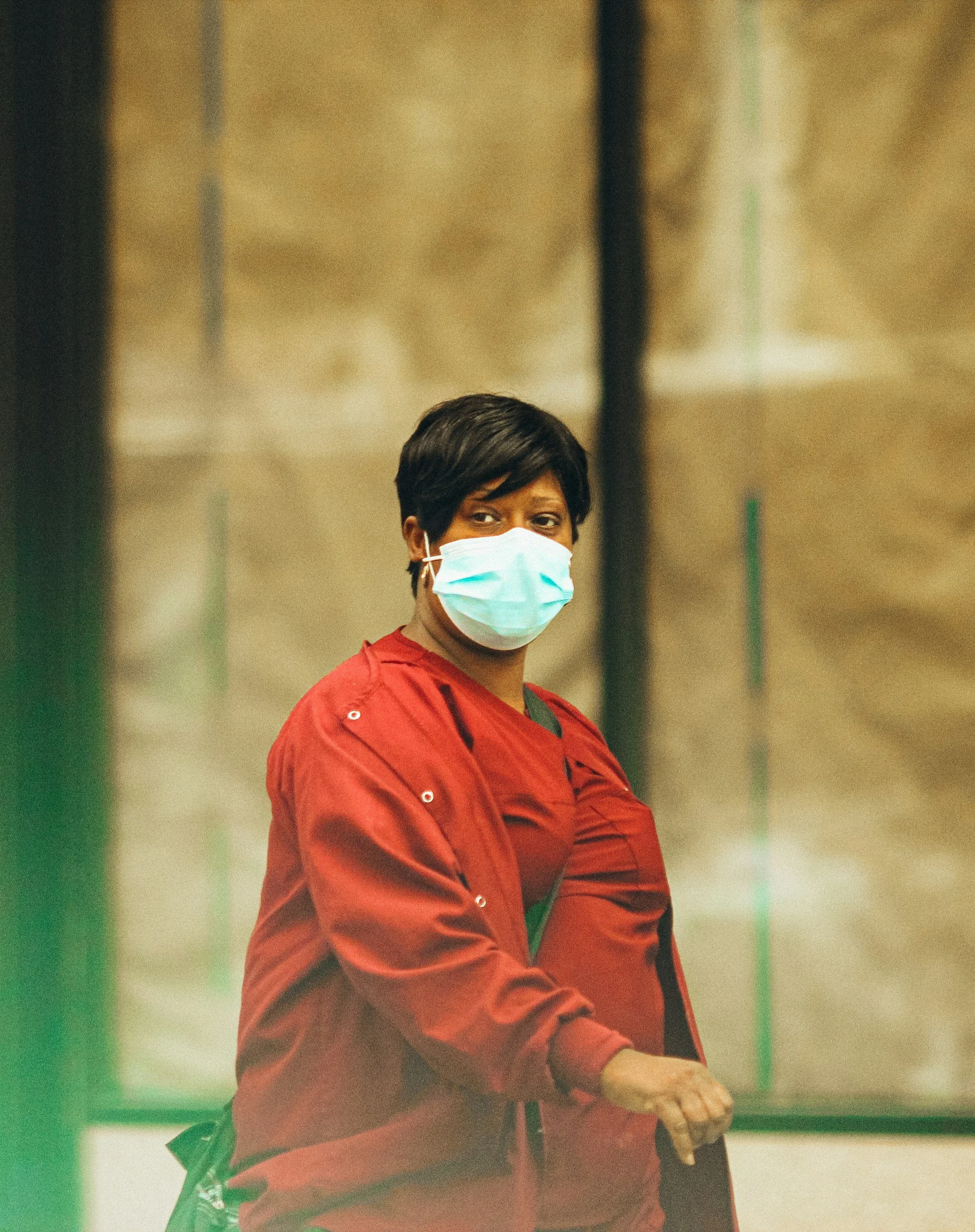 A woman wearing a red jacket and a blue surgical mask looks over her shoulder indoors against a blurred background.