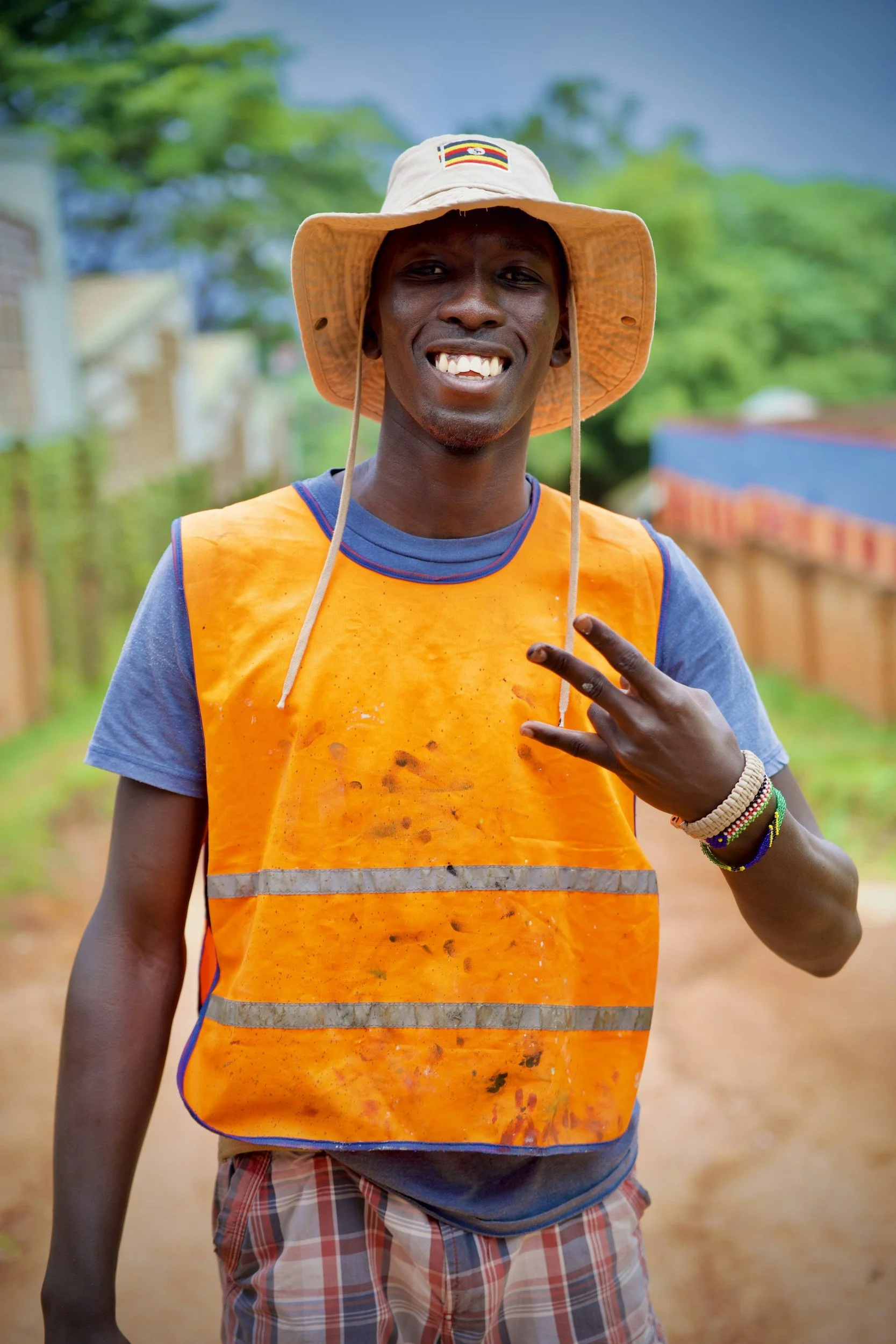 A smiling man wearing a hat and orange safety vest making a peace sign with his right hand outdoors.