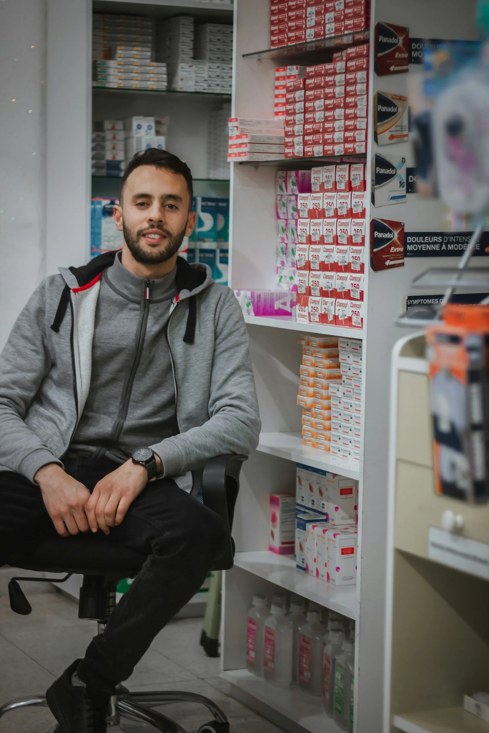 A man sitting on a rolling chair in front of pharmacy shelves stocked with various medication boxes and bottles.