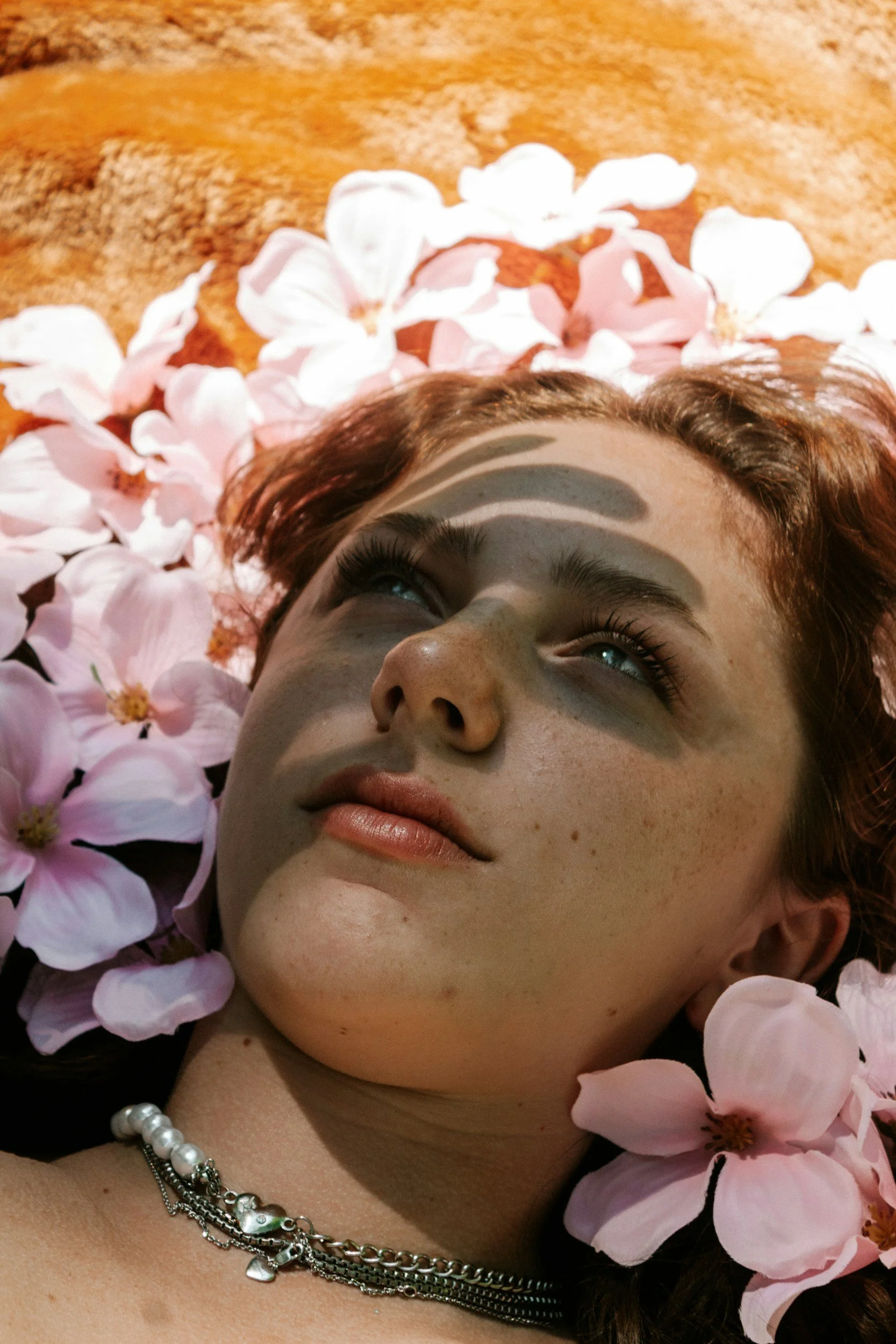 Close-up of a woman with freckles lying among pink flower petals on a sandy ground, looking upward with shadows cast on her face, wearing layered silver necklaces.