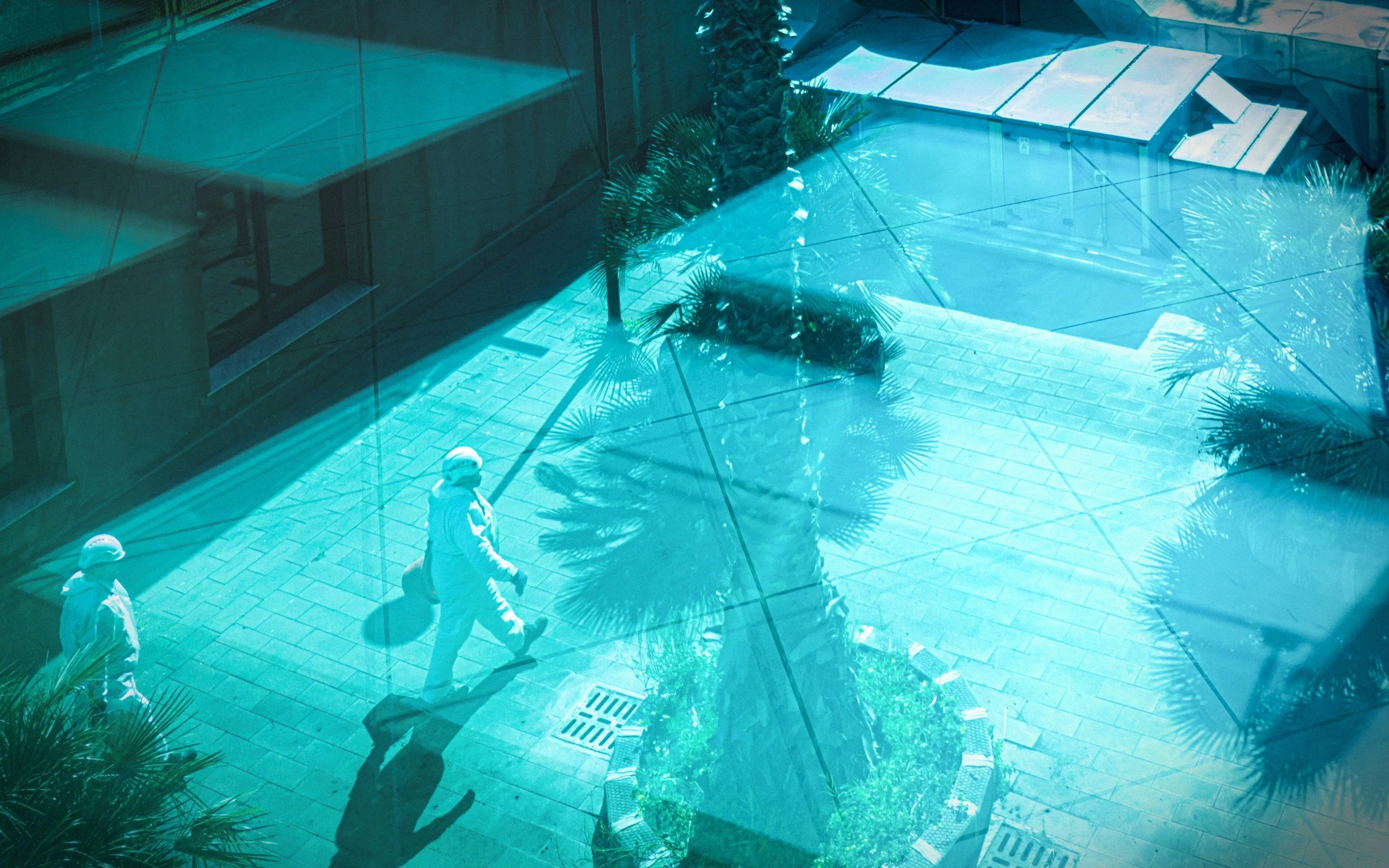 View through a reflective glass window showing two workers in helmets walking on a tiled patio near some palm trees.