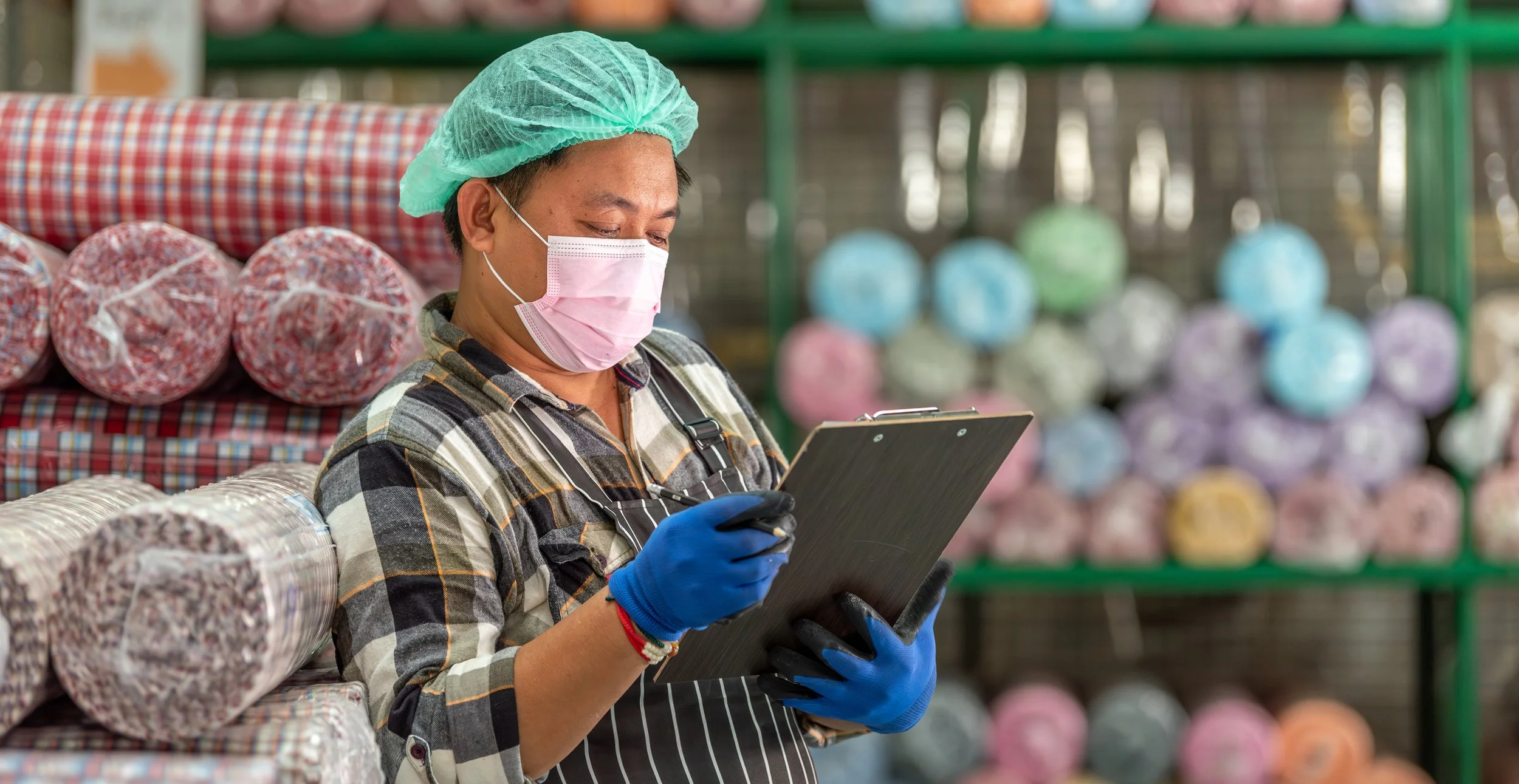 A worker wearing a face mask, hair net, and gloves, checking on inventory or orders in a store with shelves of colorful yarn in the background.