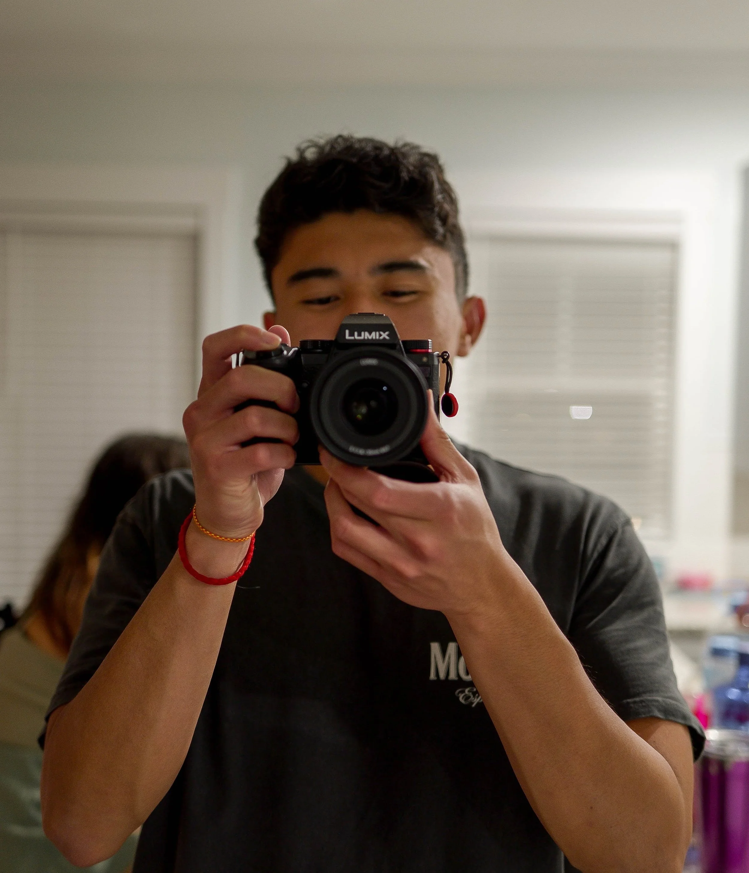 Young man taking a selfie with a camera in a room with blinds on the windows.