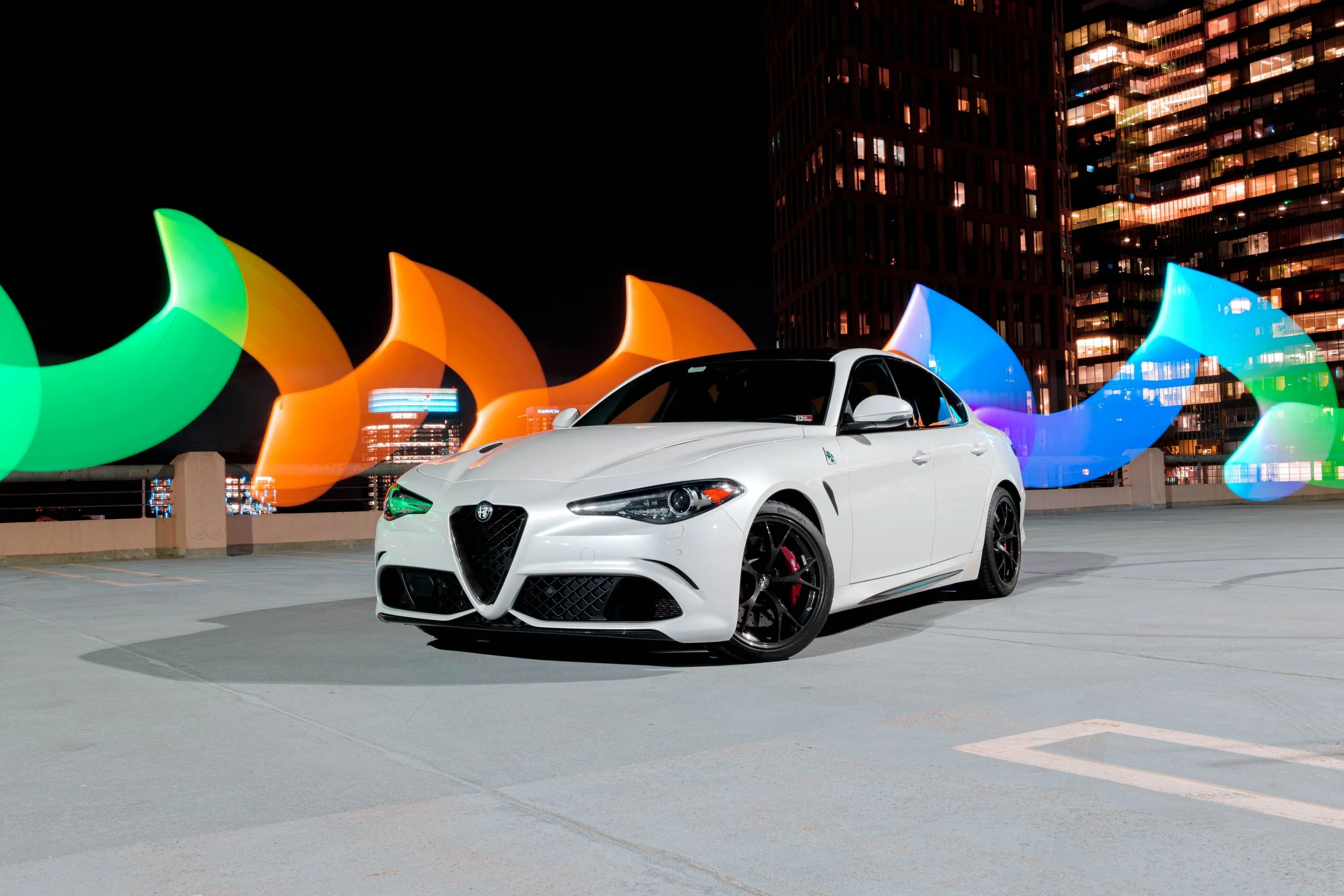 White sports car parked on a rooftop parking lot at night with colorful light trails and city buildings in the background.