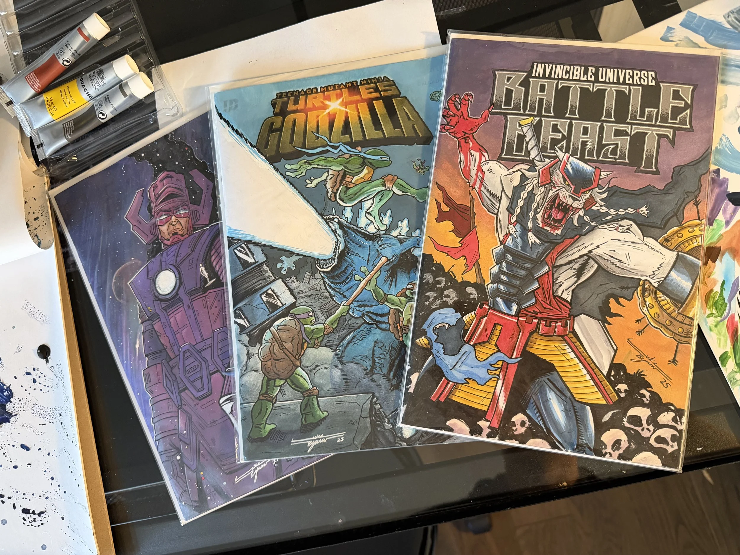 Three comic book covers featuring classic characters from Teenage Mutant Ninja Turtles, Godzilla, and Battle Beast, displayed on a black table with paints and art supplies nearby.