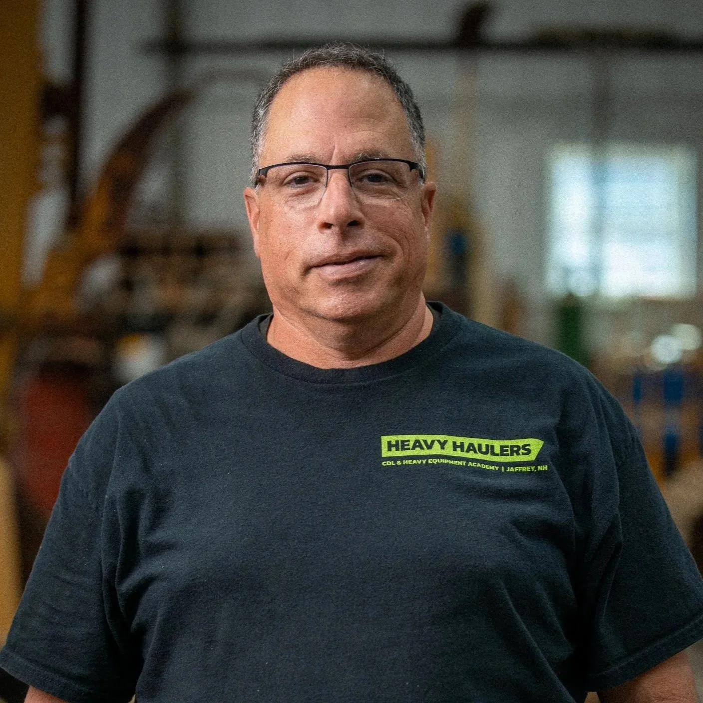 Smiling man with glasses and short hair, wearing a black t-shirt that says "Heavy Haulers" in green, standing in a workshop with equipment and machinery in the background.
