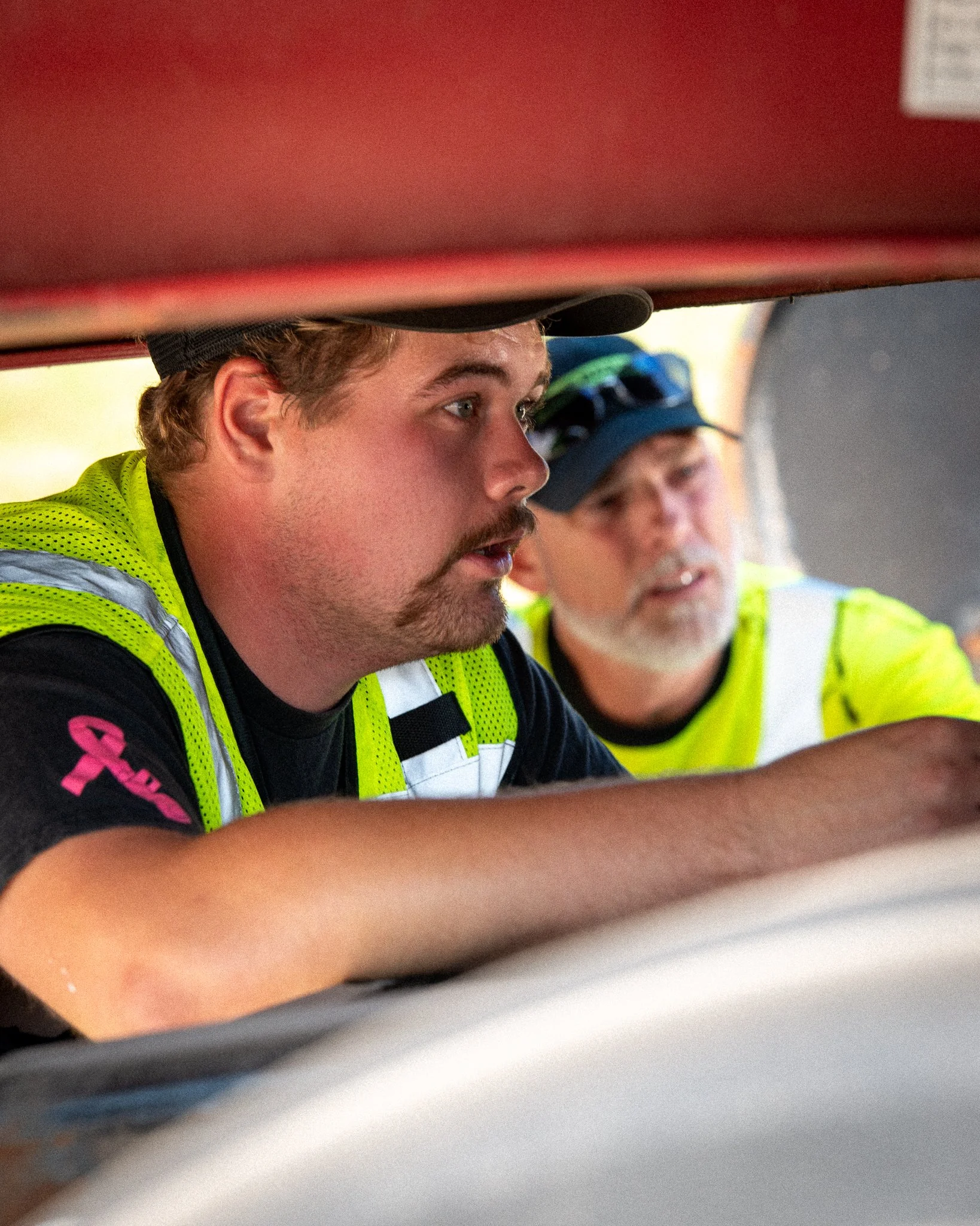 Two men wearing yellow safety vests working underneath a structure, closely examining something.