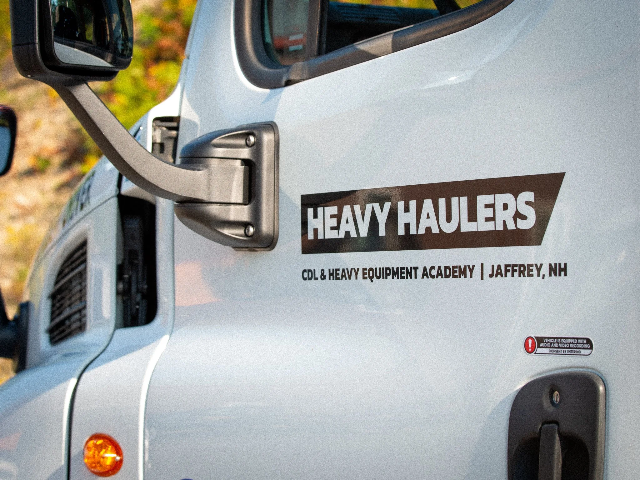 Close-up of a white heavy haulers truck with a black and white logo on the side that reads "Heavy Haulers" and below it says "CDL & Heavy Equipment Academy | Jaffrey, NH." The truck's side mirror, part of the cab, and a small portion of the front are visible, set against a background of trees with fall foliage.