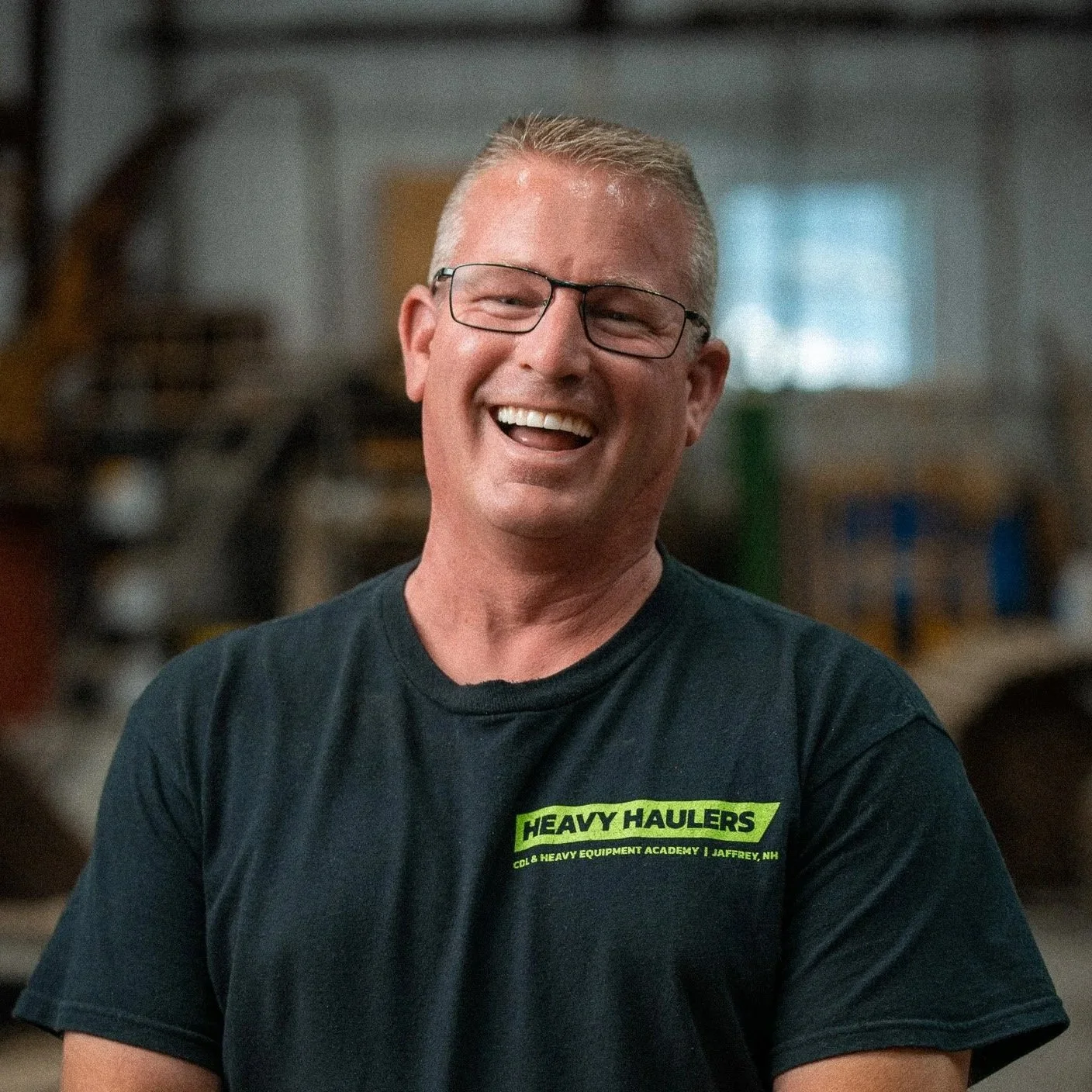Smiling man with glasses wearing a black 'Heavy Haulers' t-shirt in a workshop setting.
