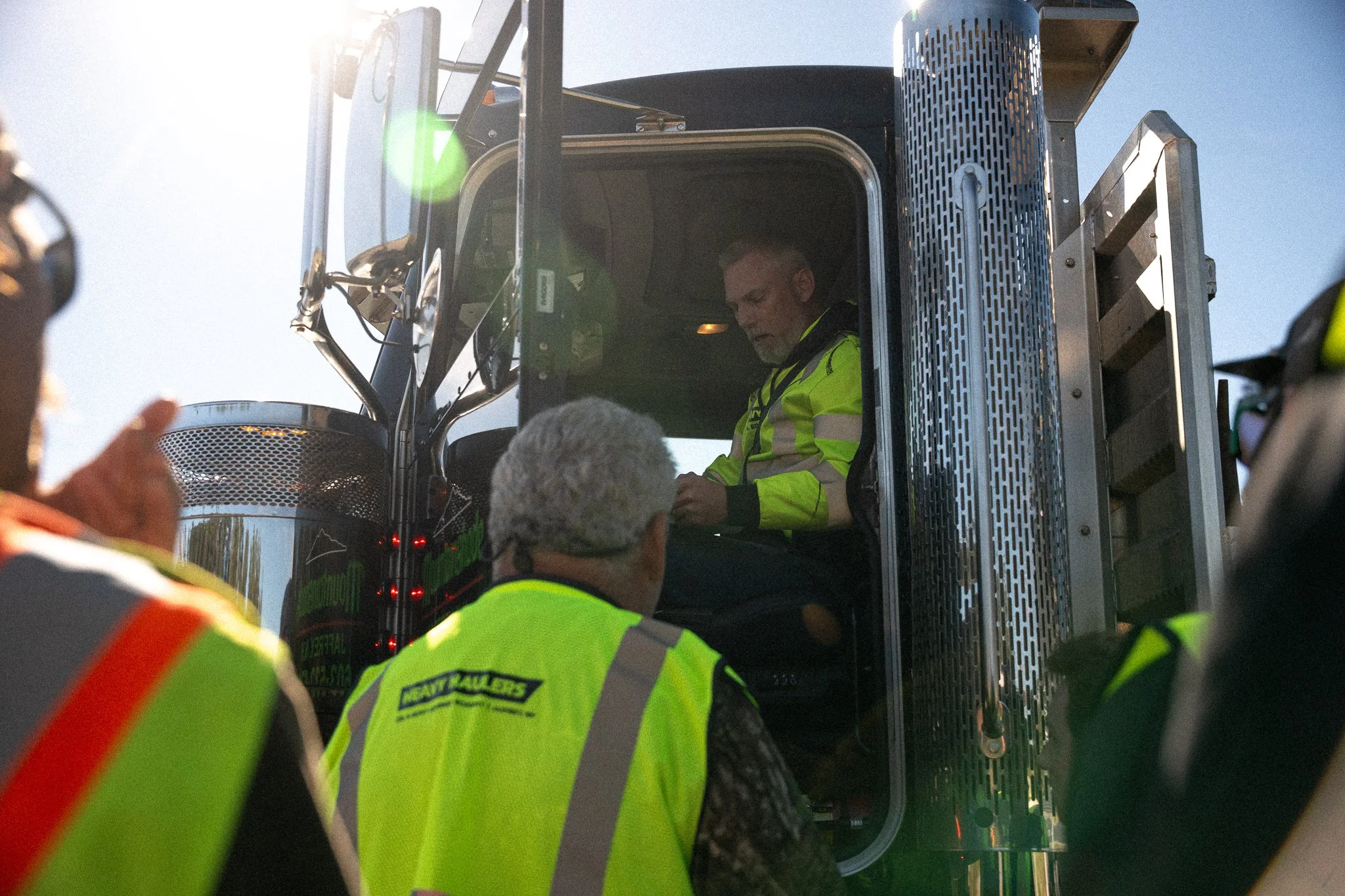 A man in a yellow high-visibility jacket sitting inside a large truck's cab, with several people outside also wearing high-visibility vests gathered around.