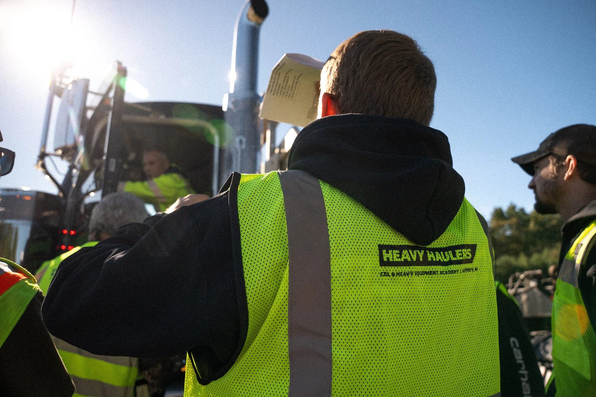 Group of construction workers wearing high-visibility safety vests, gathered outside on a sunny day, with a heavy machinery vehicle in the background.