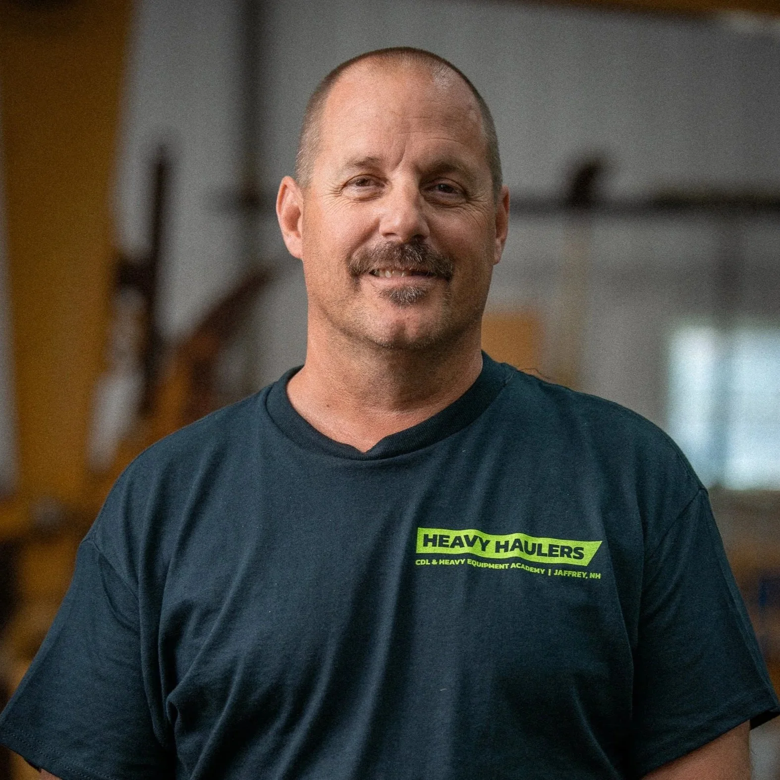 Smiling man with short hair and mustache wearing a navy blue T-shirt with bright green text that reads 'Heavy Haulers'.