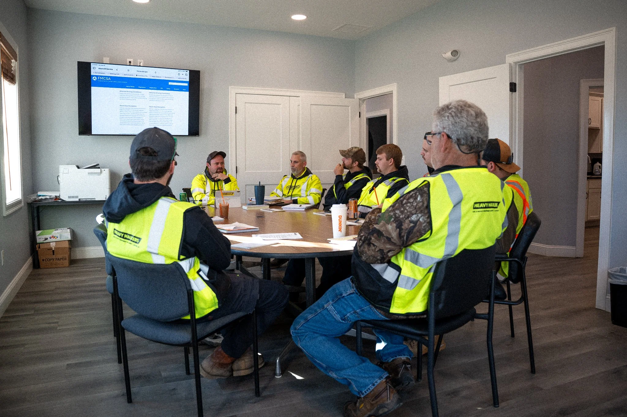 Group of construction workers in high-visibility vests gathered around a conference table in a meeting room, listening to a man speaking at the front. A large screen displays a webpage, and the room has wooden flooring, a window on the left, and an open door on the right.