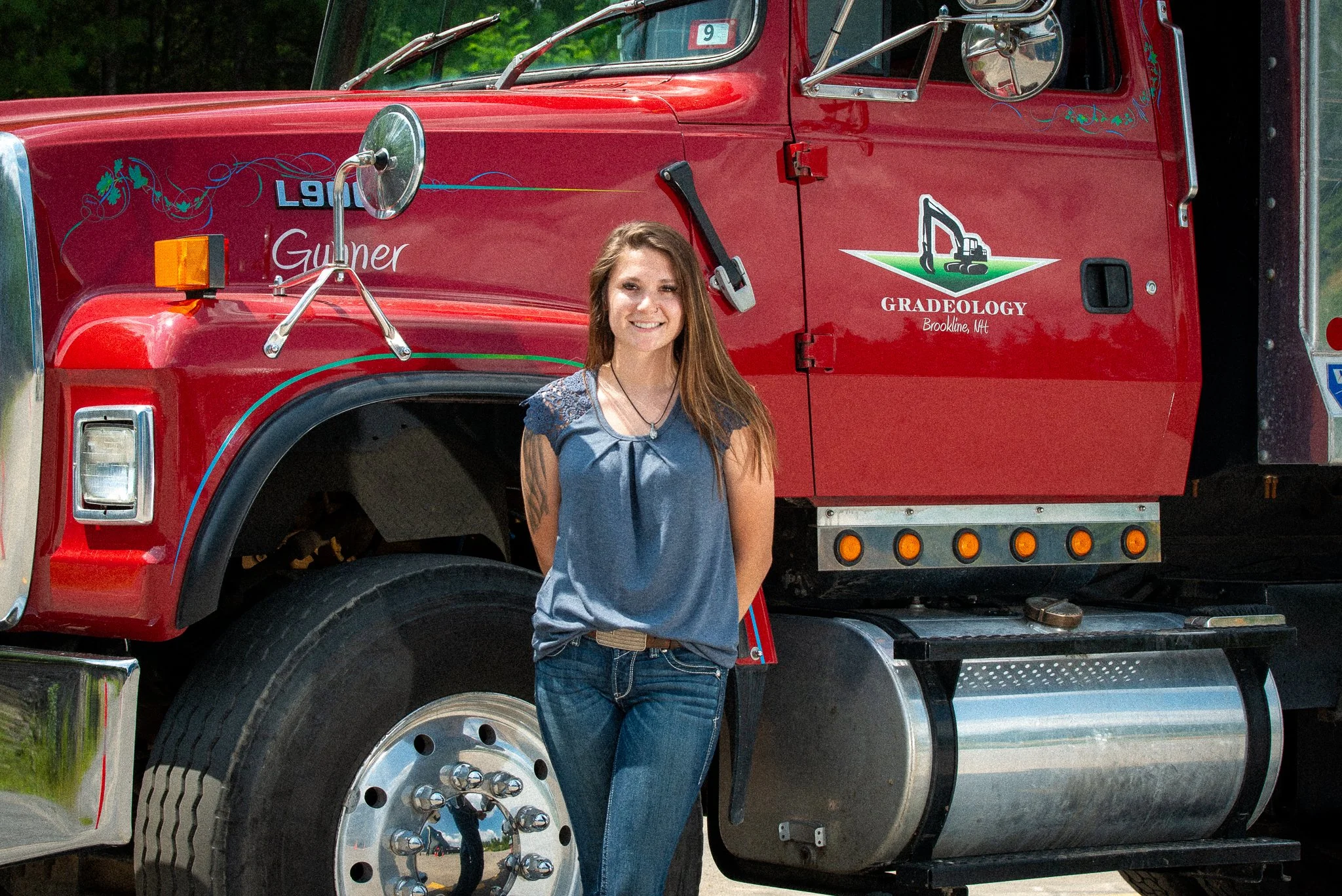 A woman smiling standing in front of a red grader truck with the logo 'Gradeology' and 'Gunner' on it, outdoors with trees in the background.
