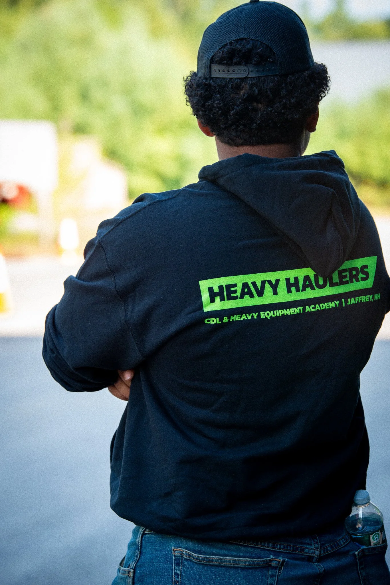 Back of a person wearing a black hoodie and a black baseball cap, with neon green text on the hoodie reading 'HEAVY HAULERS' and additional text below, standing outdoors with trees and a blurred background.