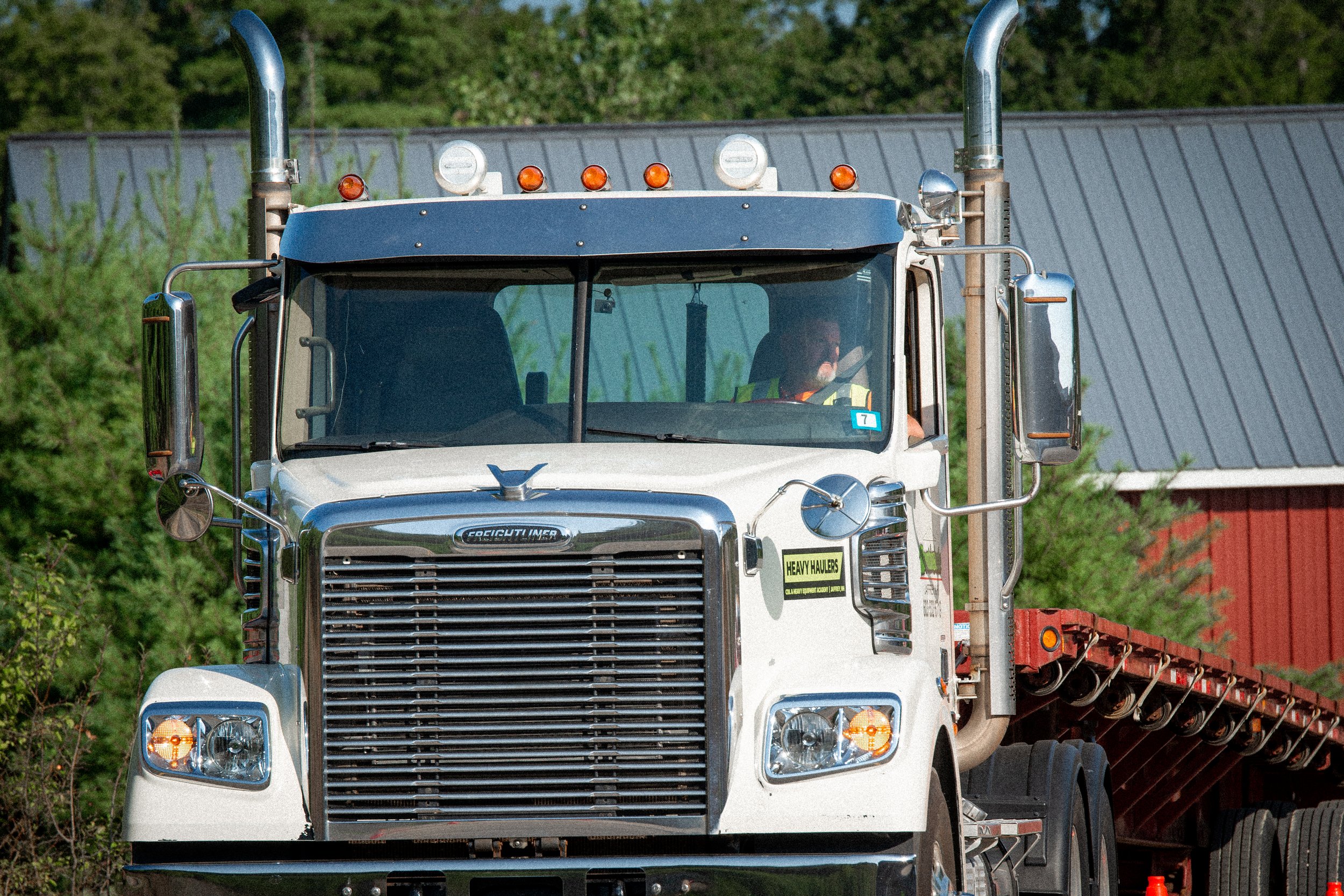 A white semi-truck driving on a rural road surrounded by green trees with red barn in the background.