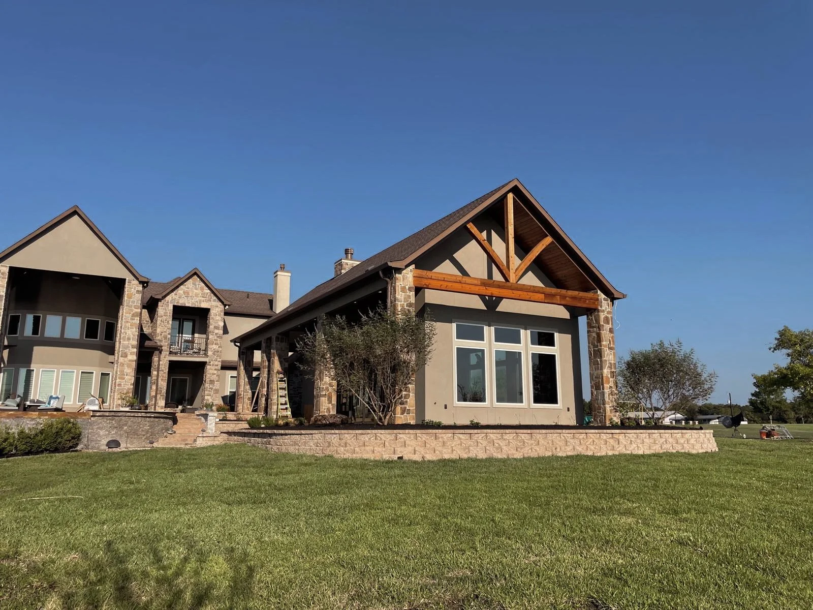 A large, modern house with stone and stucco exterior, multiple windows, and a sloped roof, next to a well-maintained lawn under a clear blue sky.