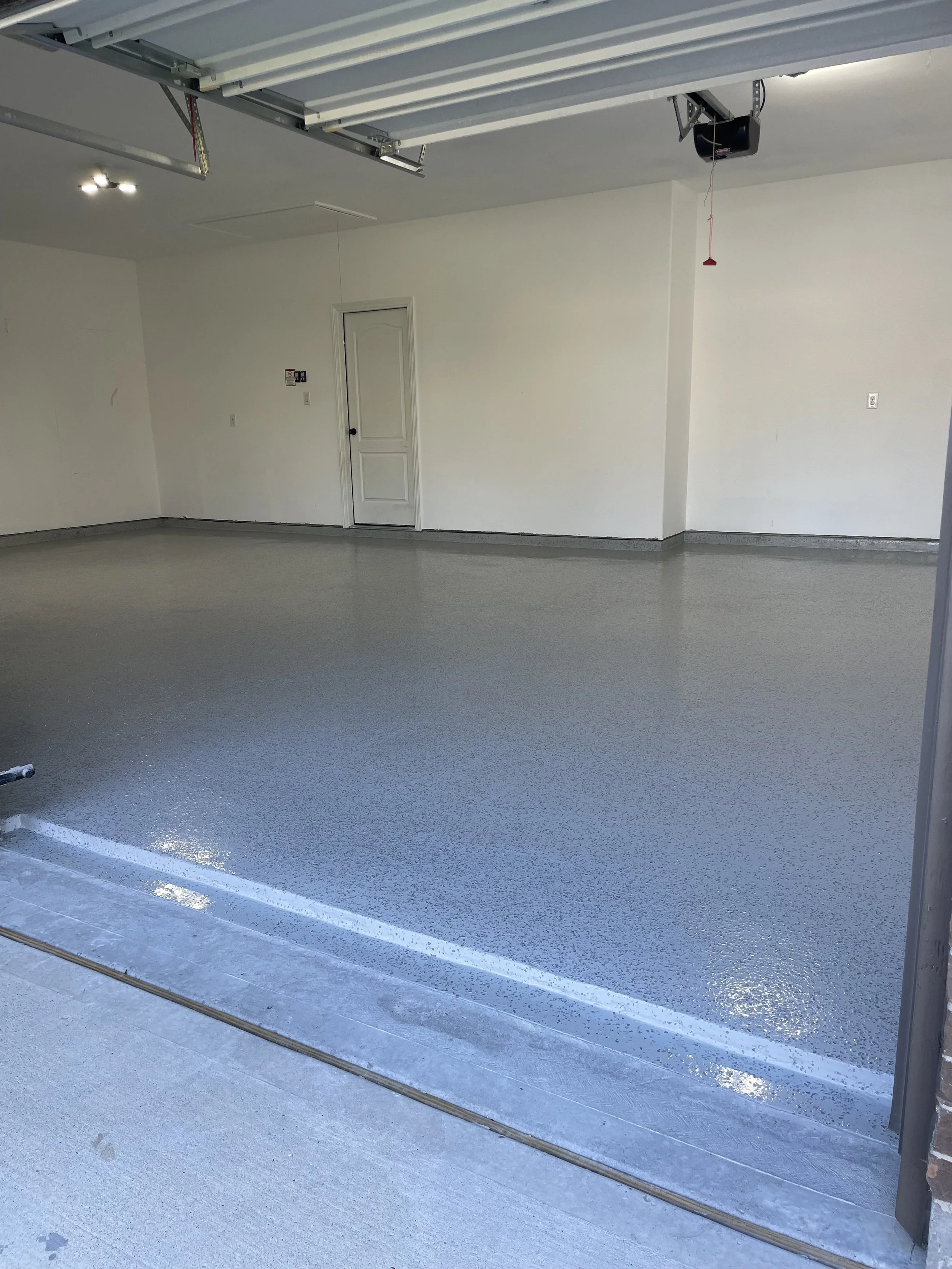 Empty garage with freshly coated epoxy floor, white walls, and a partially open garage door.