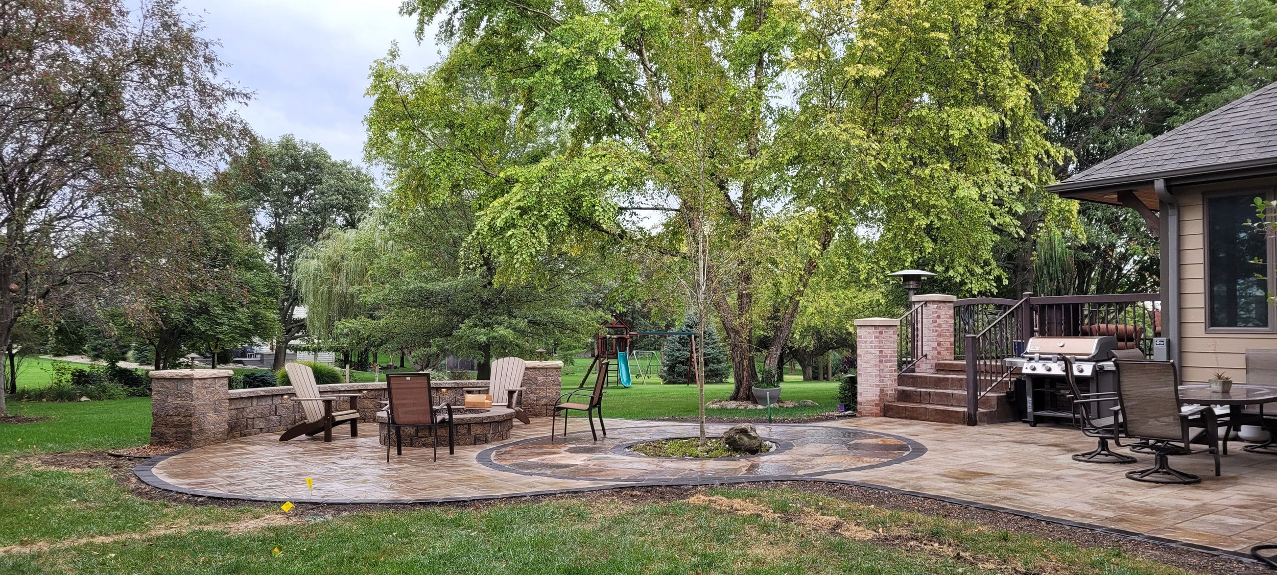 A backyard patio with stone paving, outdoor seating, a charcoal grill, a small tree, a playset, and lush green trees in the background.