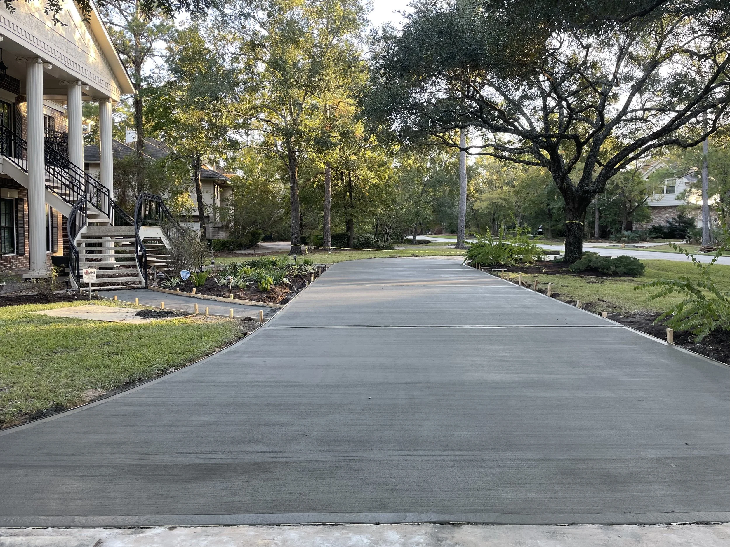 Concrete driveway leading to a residential house with stairs and columns, surrounded by a landscaped yard with trees and plants.