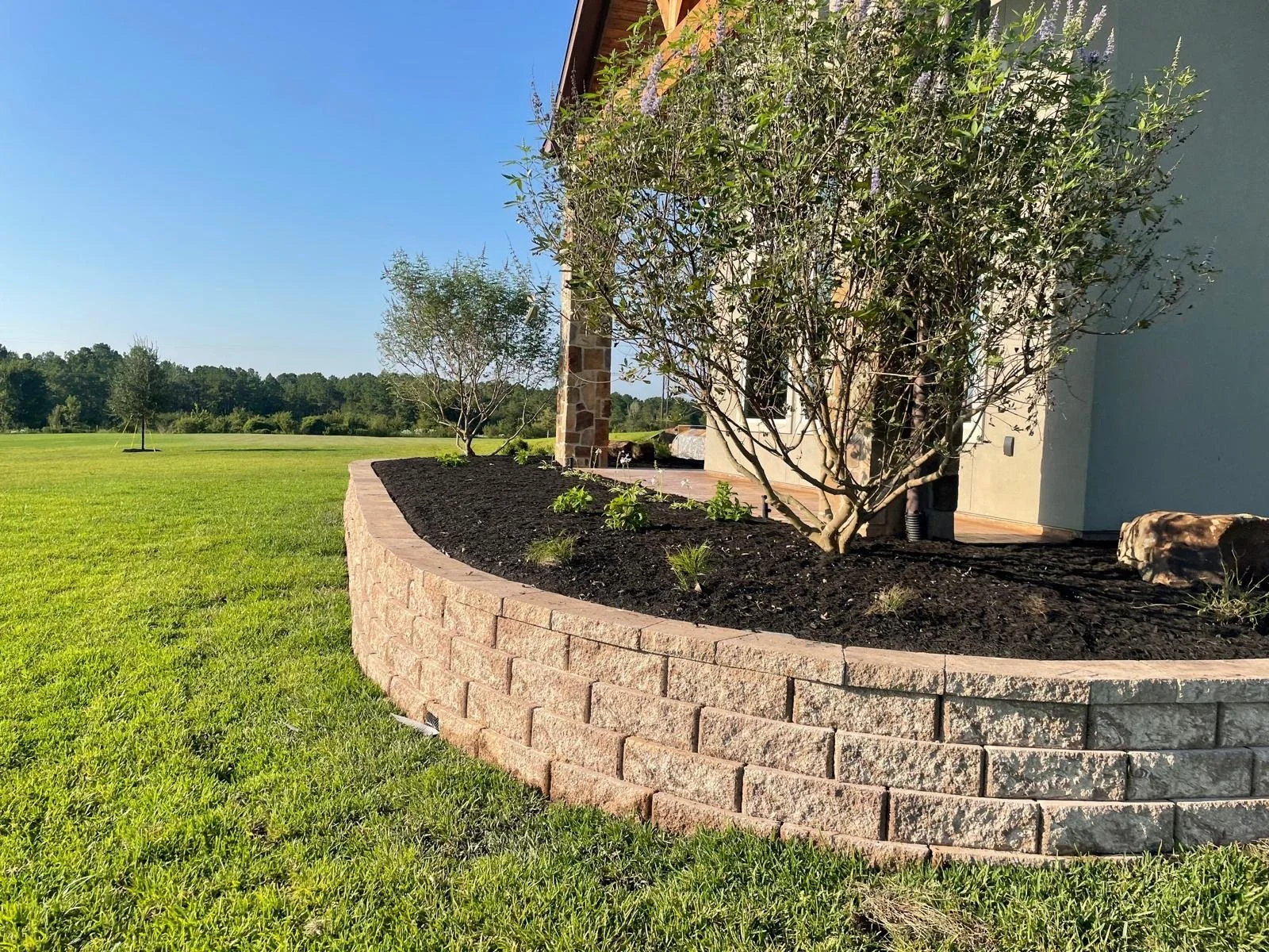 A landscaped garden bed with dark mulch and small plants, bordered by a low, curved stone wall, next to a house with a light blue exterior. The scene includes trees and a green lawn under a clear blue sky.
