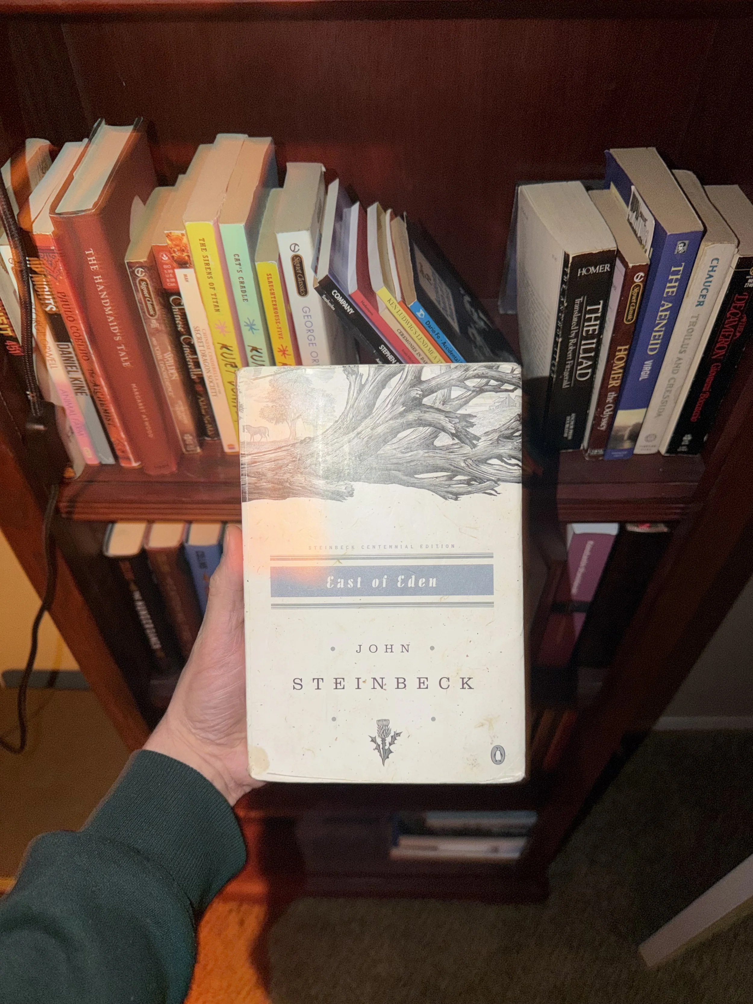 Person holding the book "East of Eden" by John Steinbeck in front of a bookshelf filled with various books.