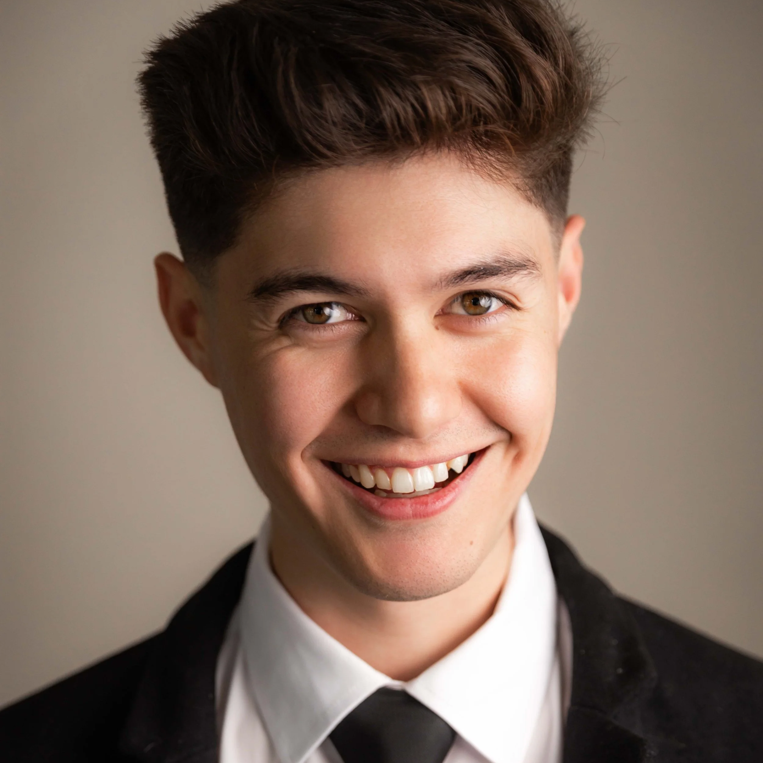 Young man with brown hair and hazel eyes, smiling, in a black suit and white shirt, gray background.