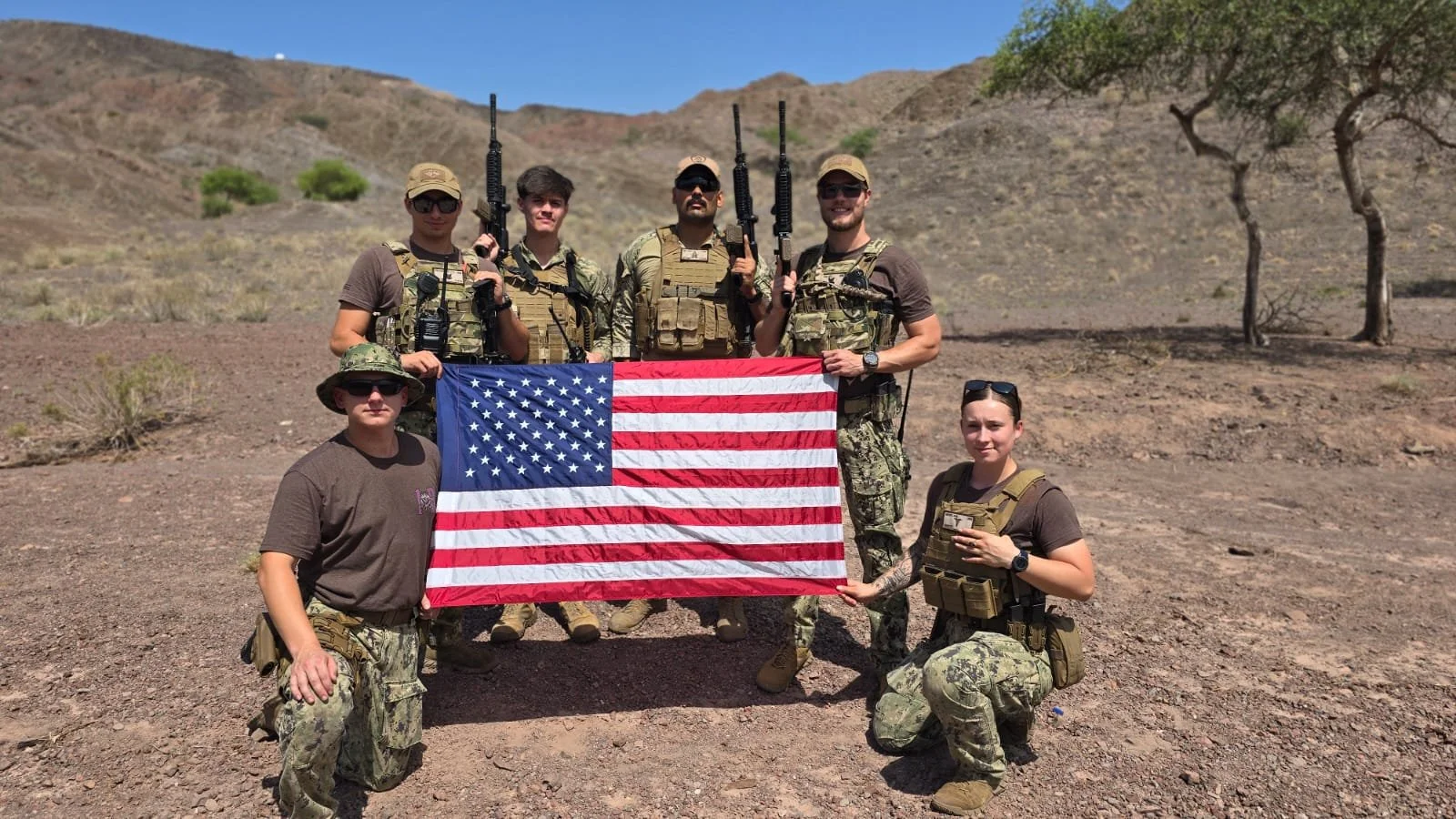 Group of six expeditionary Sailors in tactical gear holding an American flag in a desert landscape with hills and sparse trees.