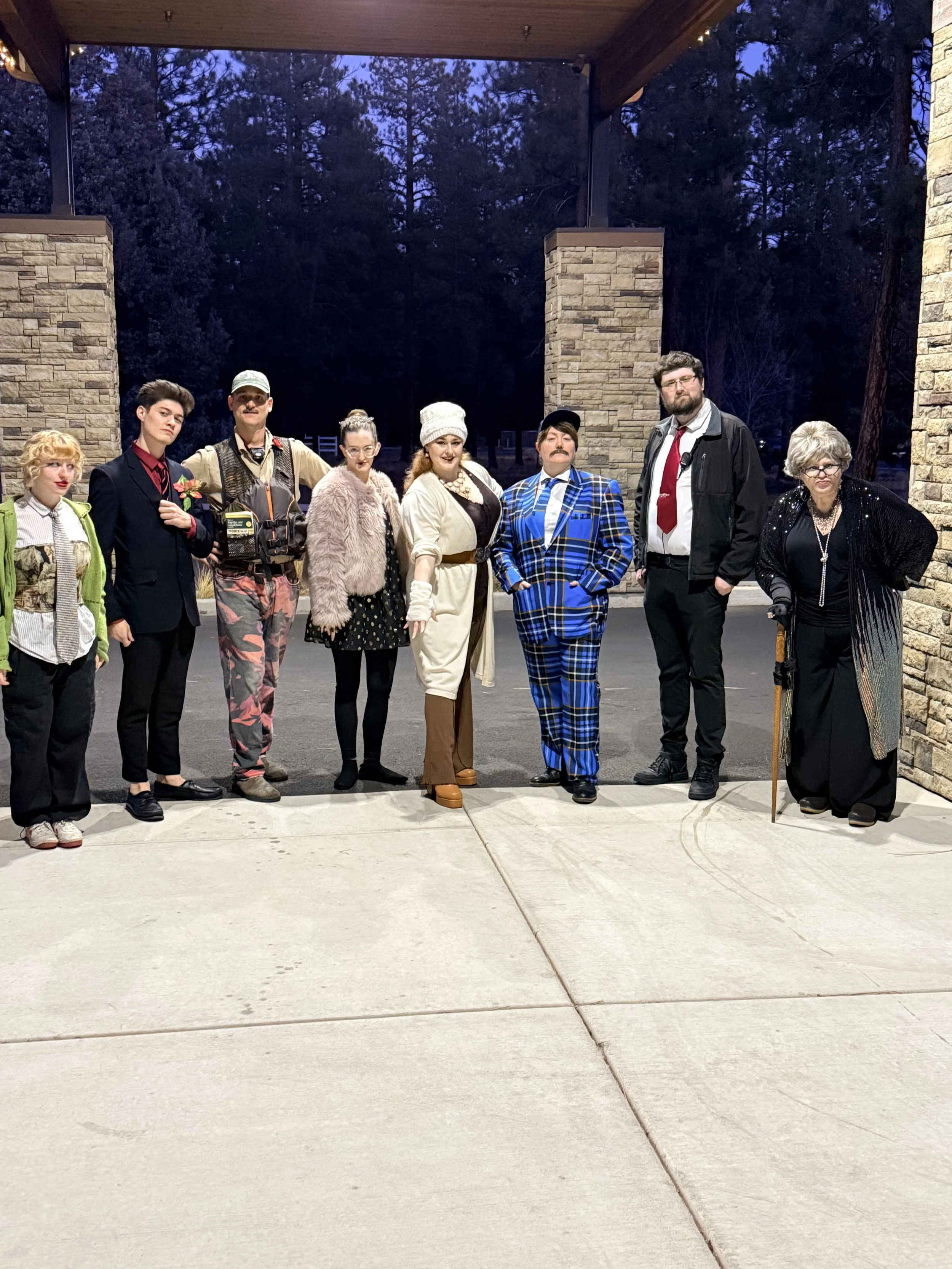 A group of eight people dressed in various costumes standing outdoors at night with a stone wall and dark trees in the background.