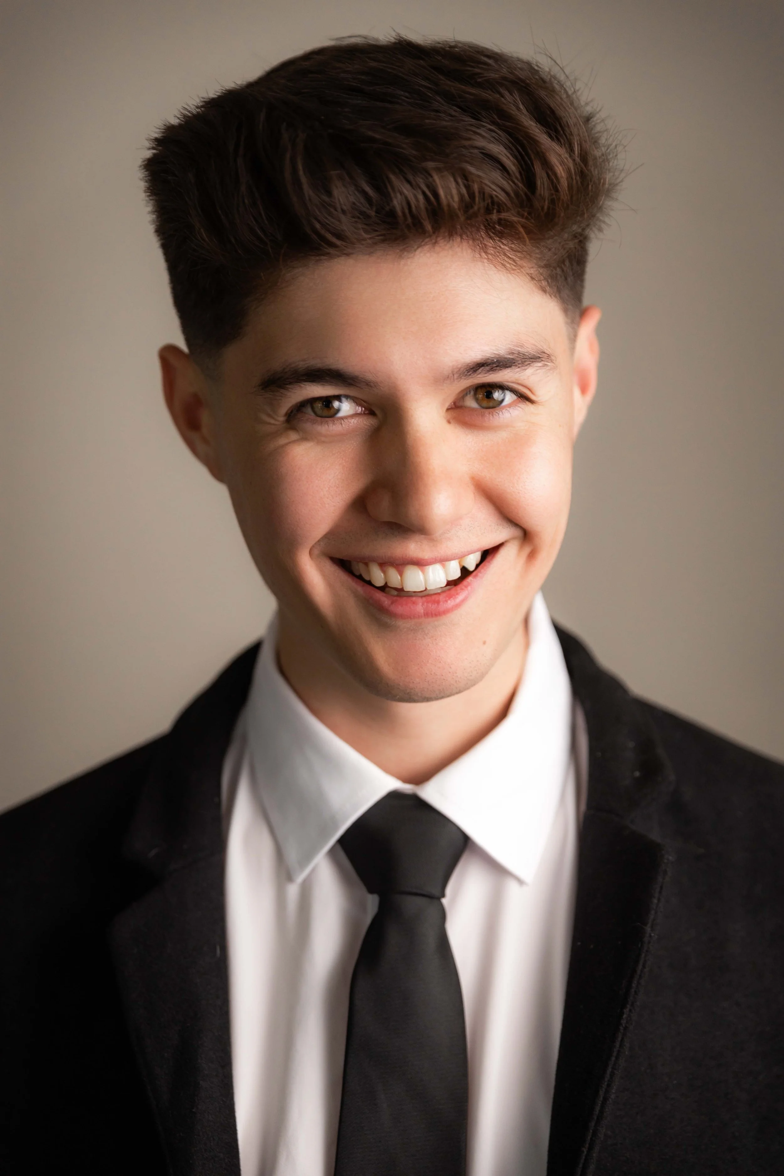 A photo of Kim, a young man with brown hair and hazel eyes, smiling, dressed in a black suit, white shirt, and black tie.