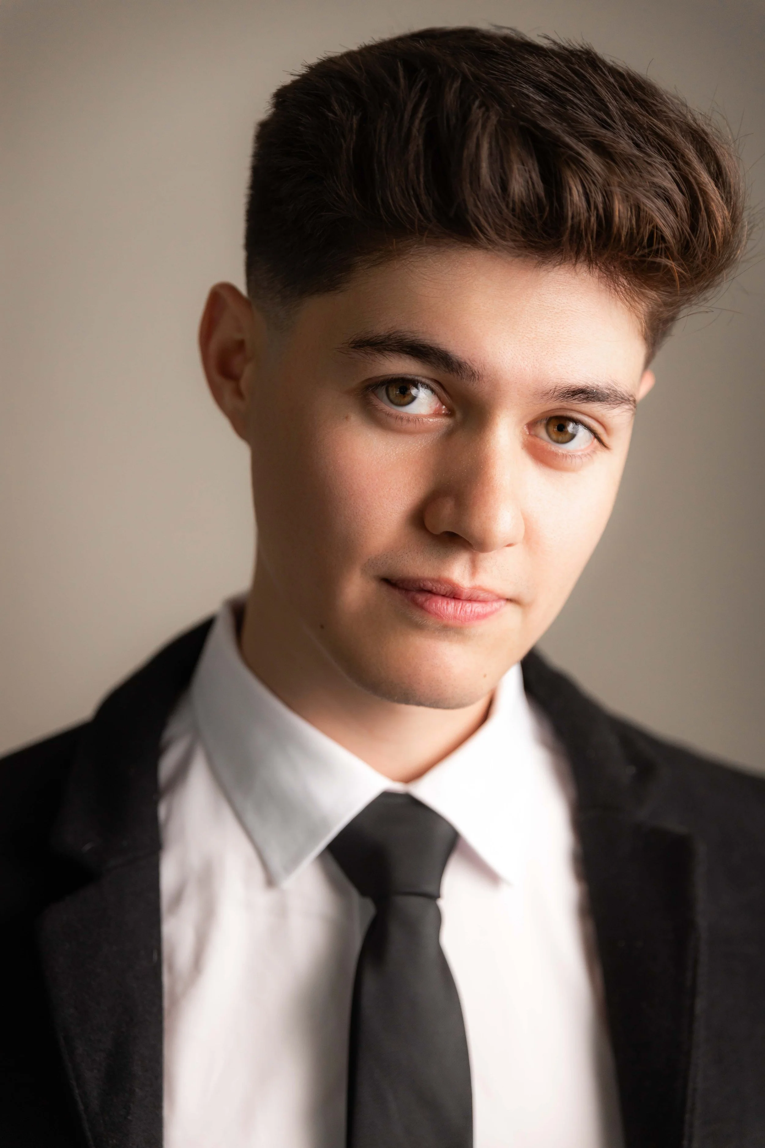 A young man with brown hair and hazel eyes, wearing a white shirt, black tie, and black blazer. He's looking at the camera with a slight smile.