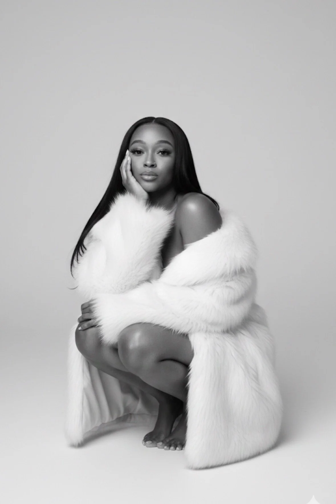 Black and white photo of a woman crouching, wearing a luxurious white fur coat. She has long straight hair and looks directly at the camera with a neutral expression, one hand resting on her face.
