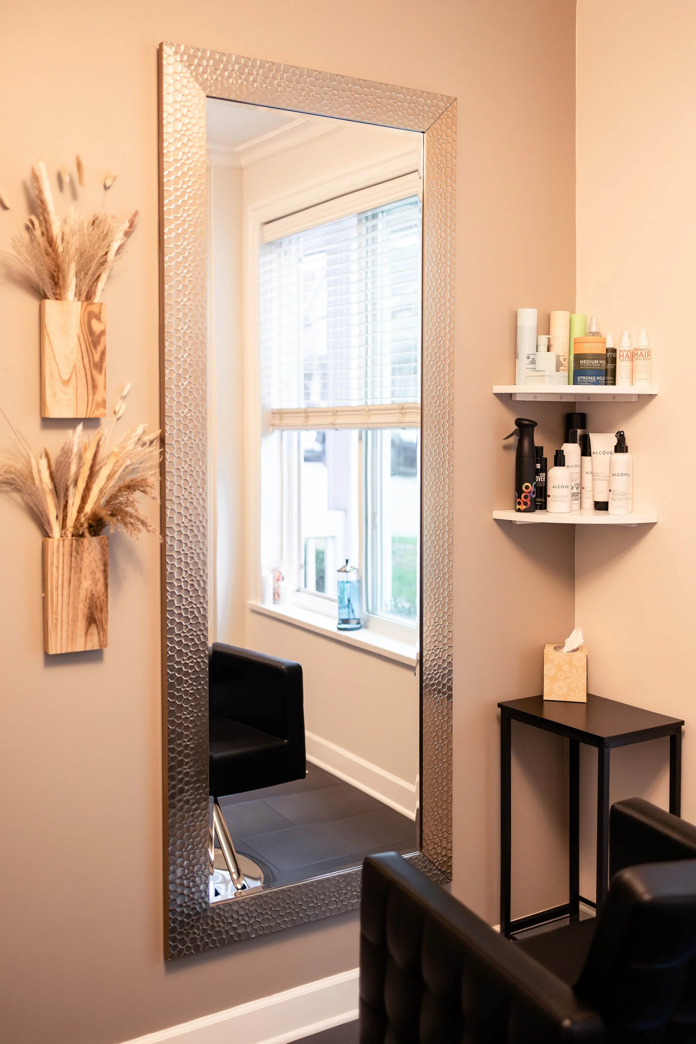 A salon or beauty room with a large decorative mirror on a beige wall. Next to the mirror, there are two wooden shelf boxes with dried pampas grass. On the right, there are two white wall-mounted shelves holding various hair and skincare products. Be