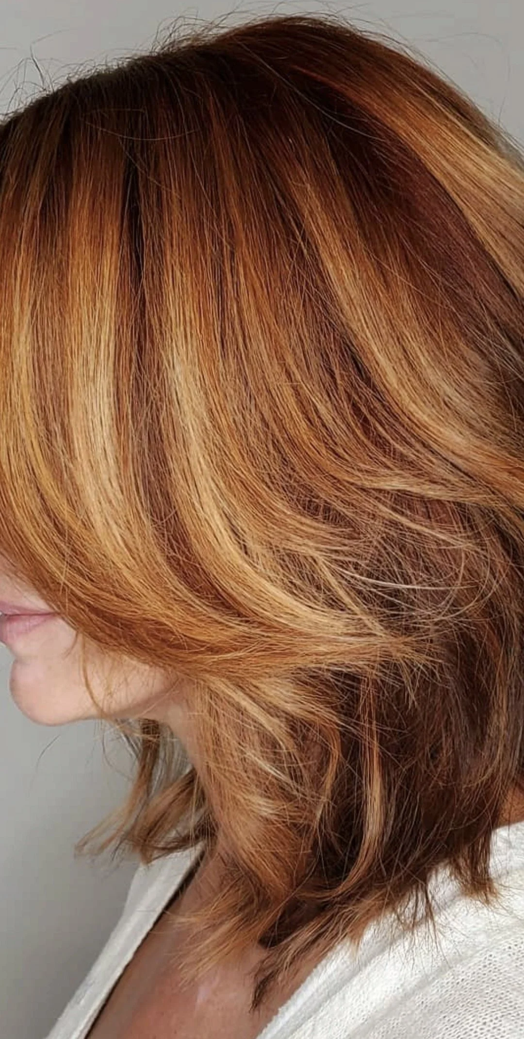 Close-up of a woman's multi-tonal copper red hair styled in loose waves, with part of her face and shoulder visible, against a plain background.