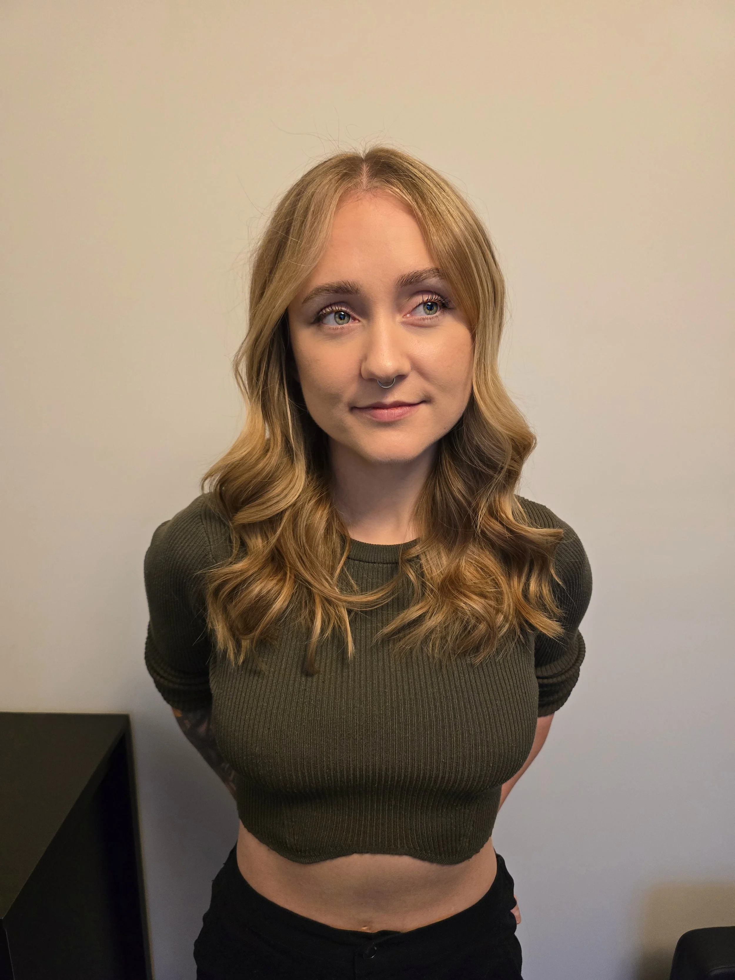 A young woman with wavy, blonde hair and a septum piercing, standing with her hands behind her back against a wall.