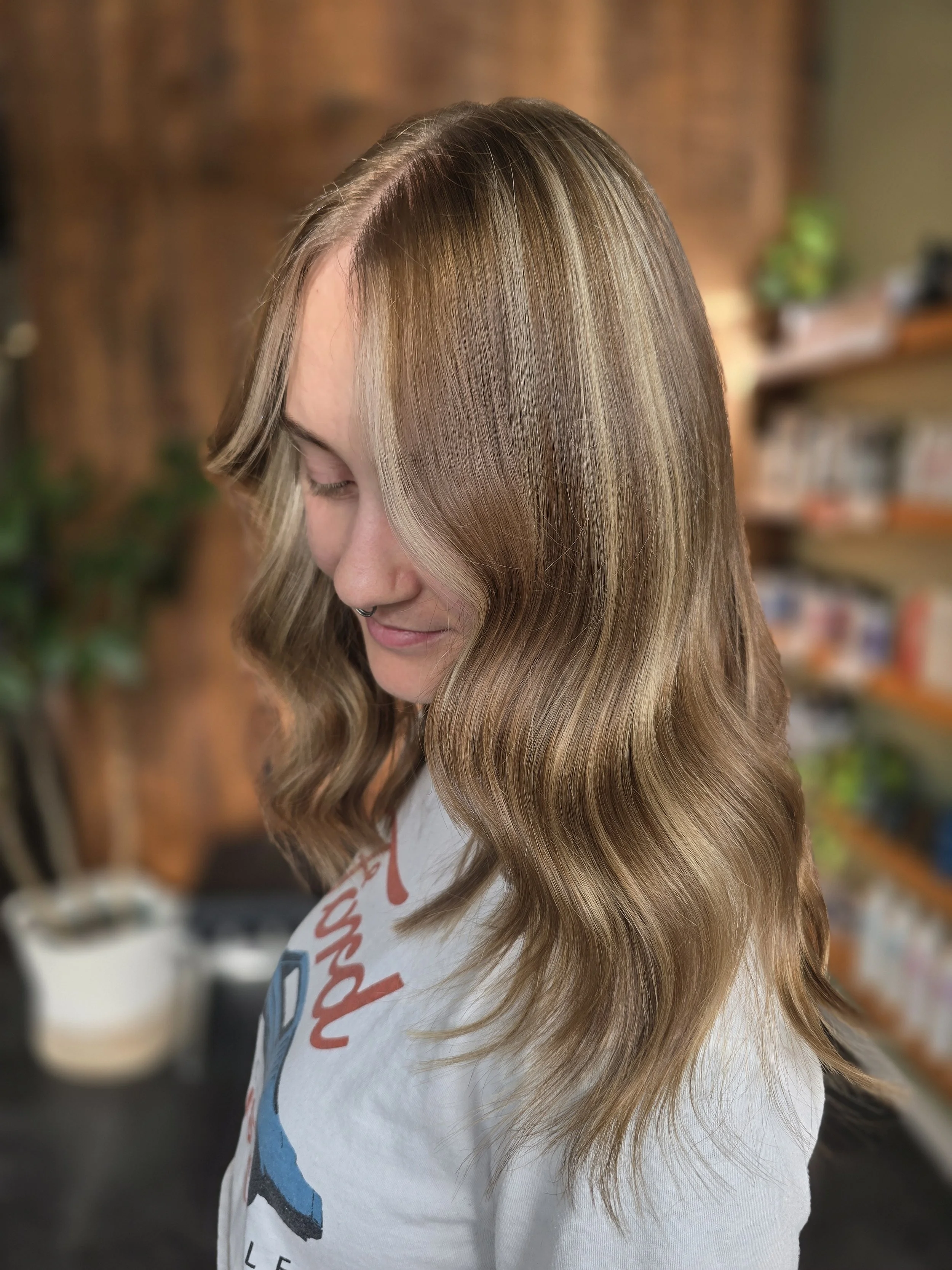 Close-up of a woman with long, wavy, light brown hair with blonde highlights, wearing a white t-shirt with a colorful graphic, smiling with her eyes closed in an indoor setting with blurred background.