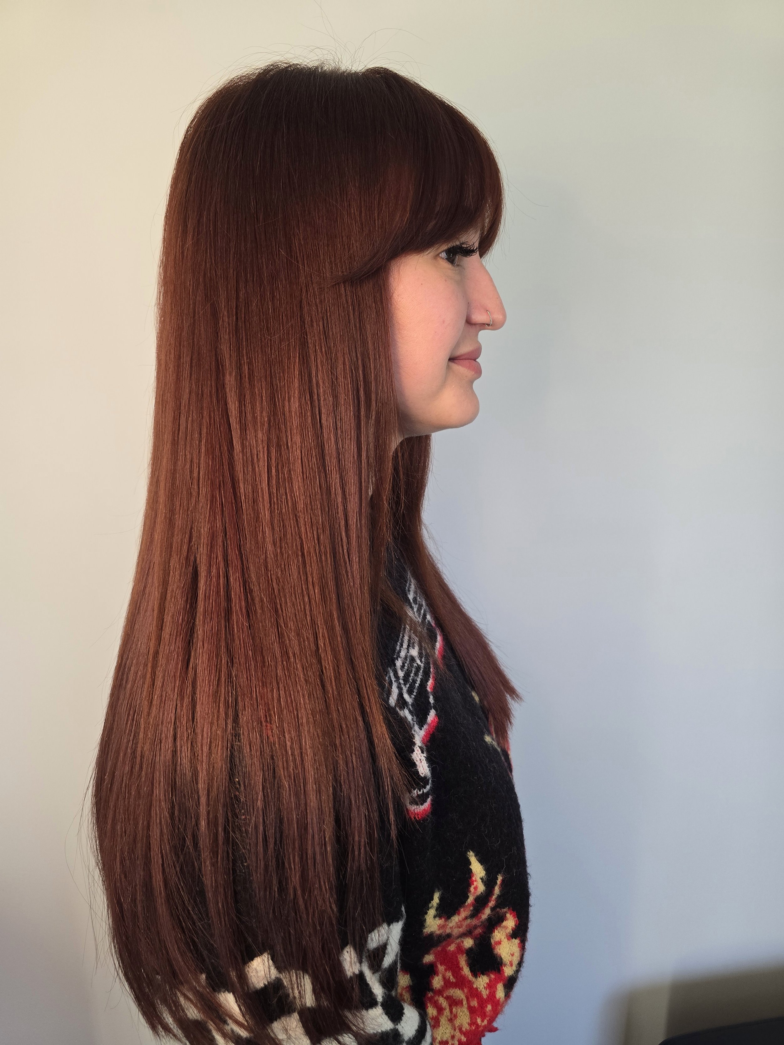 Profile side view of a woman with long, straight, deep copper-brown hair, wearing a patterned sweater, standing against a  light-colored wall.