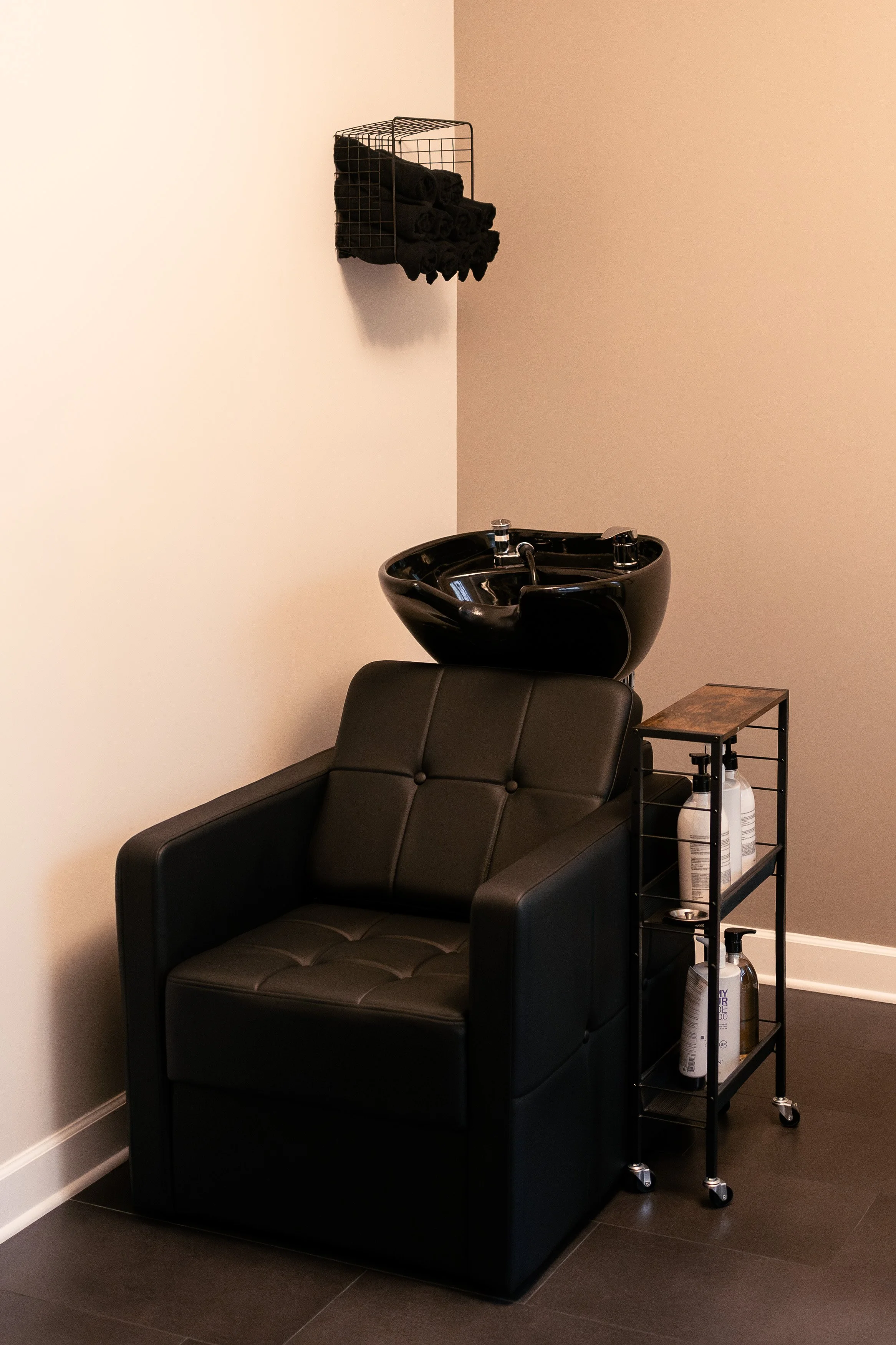 Black salon chair with a black sink for hair washing behind it, next to a cart with hair and scalp treatment products, and black towels stored in a wall-mounted rack.
