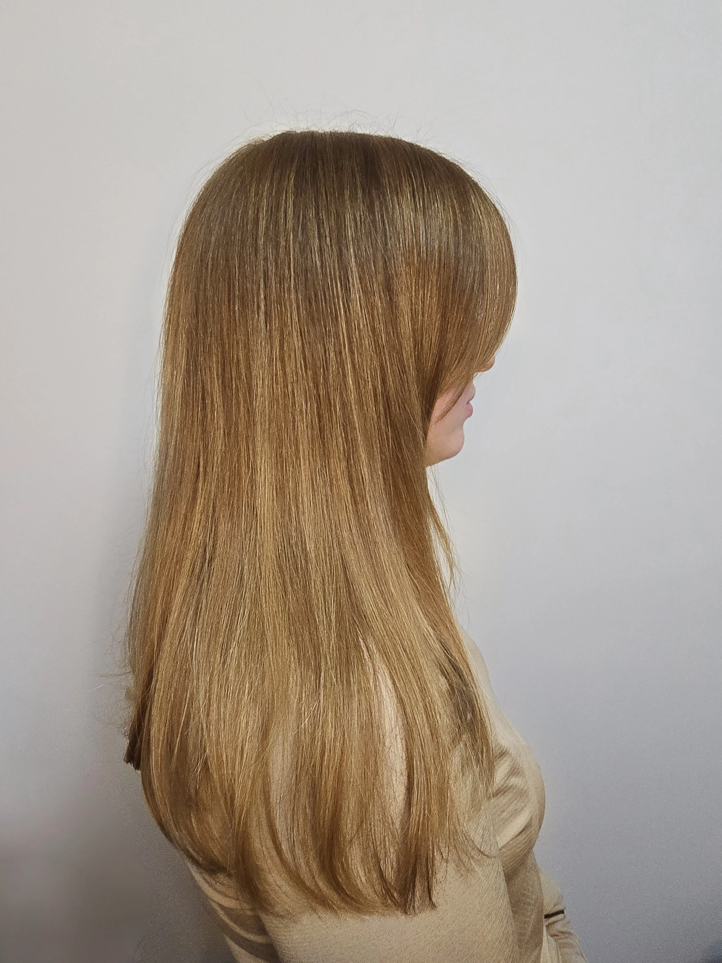 Side profile of a woman with long, straight, light brown hair with golden blonde highlights wearing a beige top, standing against a white wall.