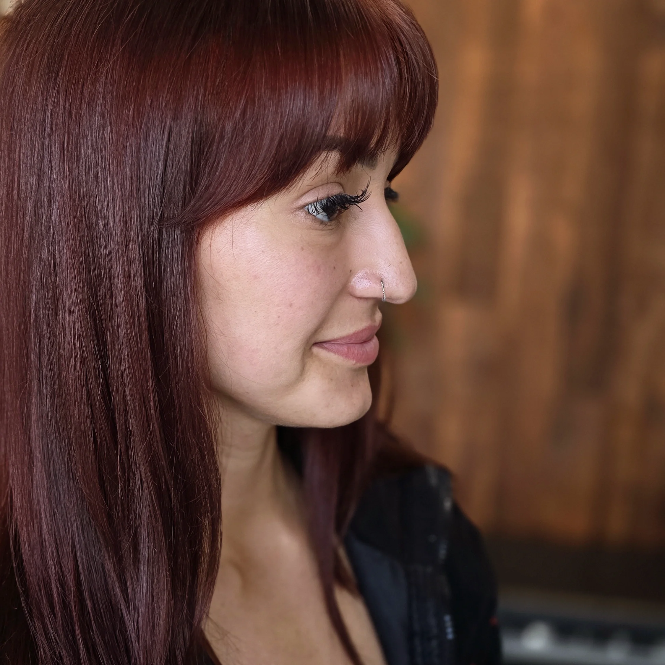 Close-up side profile of a woman with long reddish-brown hair and a nose piercing, wearing black clothing, against a blurred brown background.