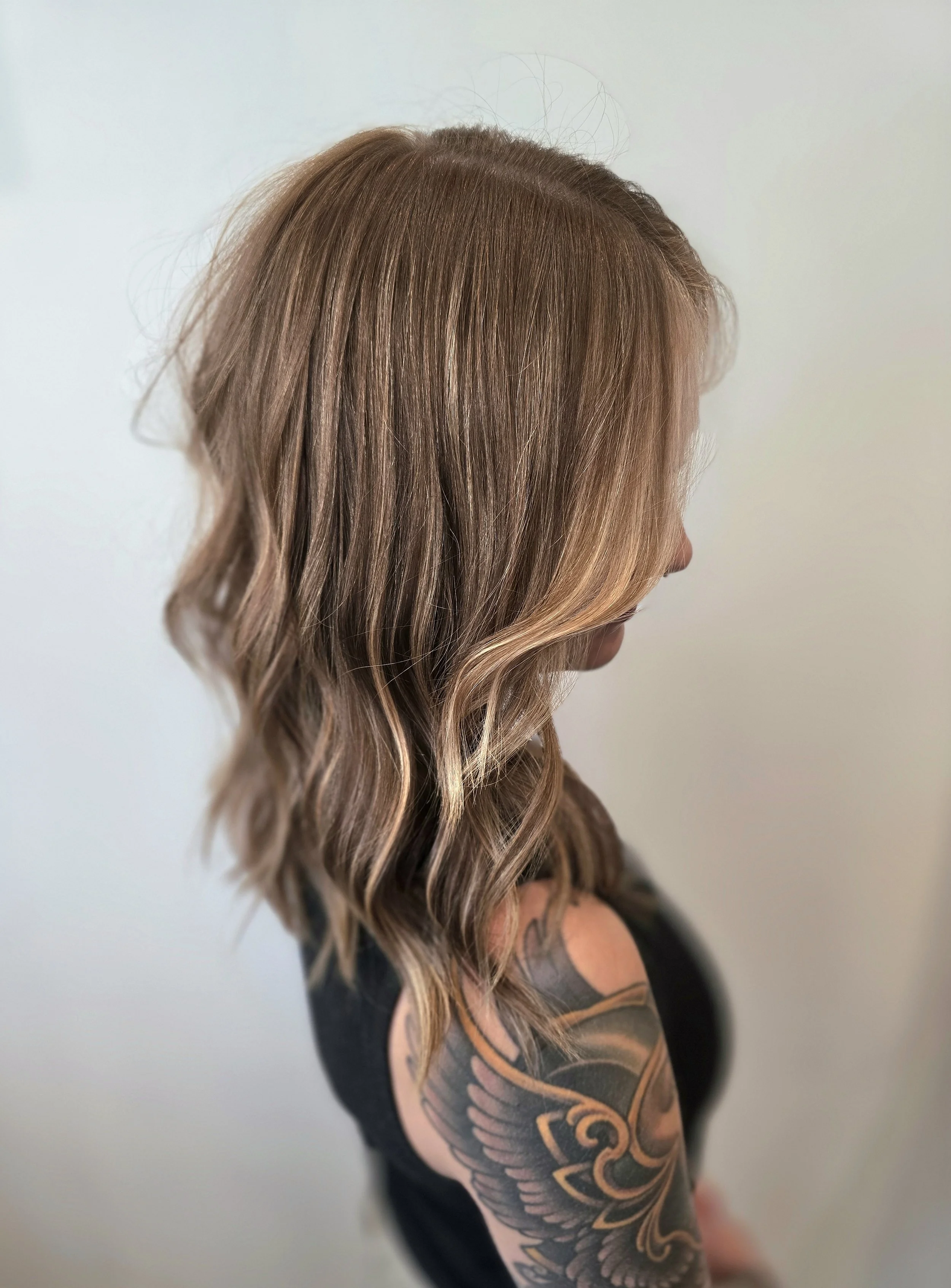 Side view of a woman with shoulder-length wavy ashy blonde hair, wearing a black tank top, revealing a colorful tattoo on her shoulder and upper arm.