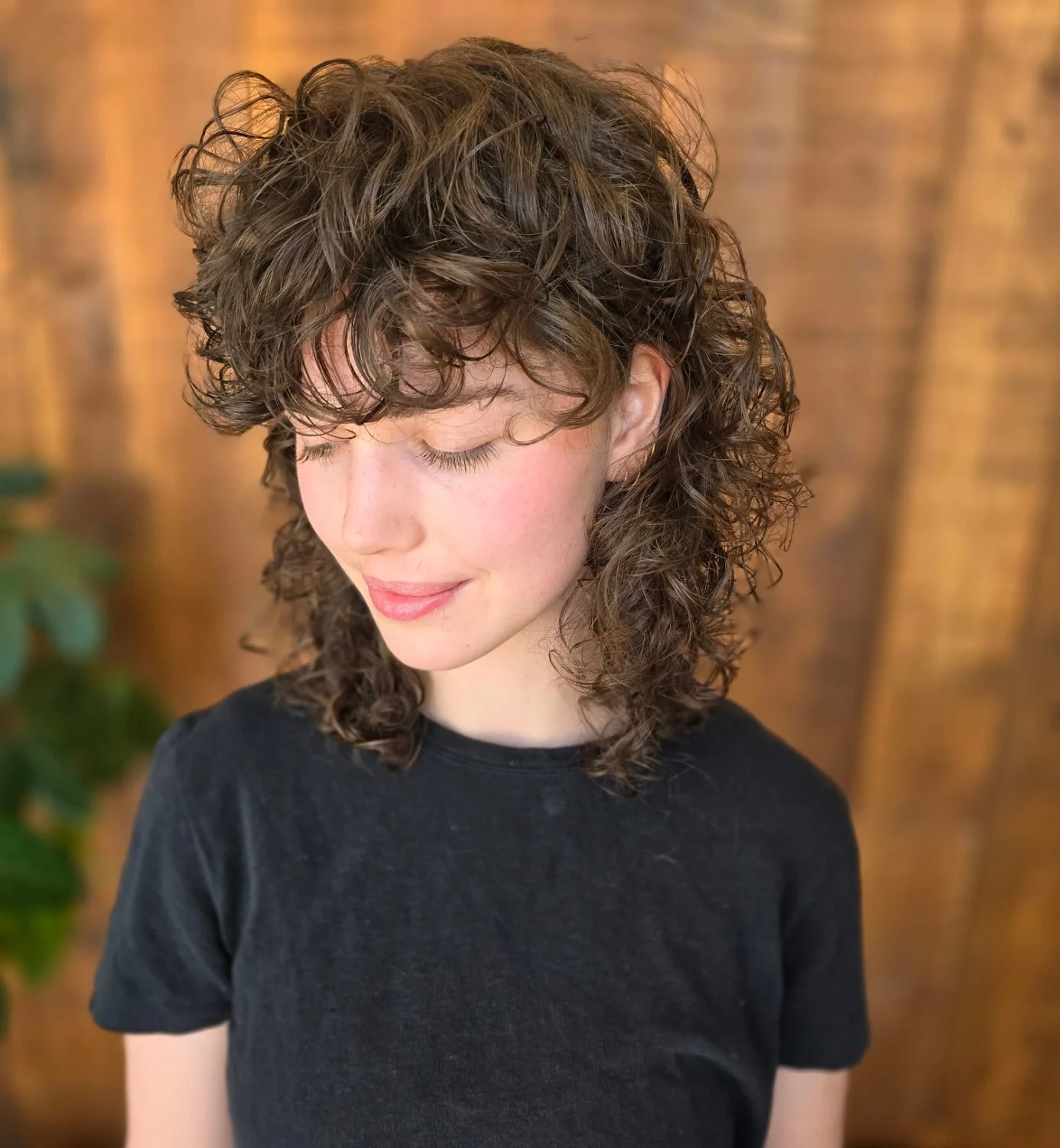 A young woman with curly brown hair, cut into an 80's inspired curly shag, wearing a black shirt, looking downward with a gentle smile.