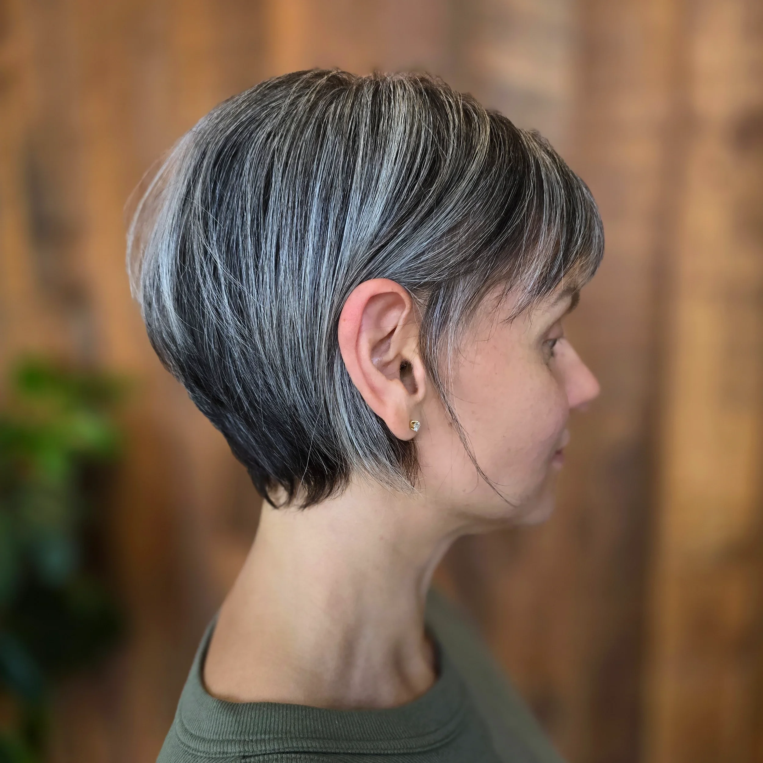 Side profile of a woman with short, beautifully coloured salt and pepper hair, wearing a small earring, against a blurred wooden background.