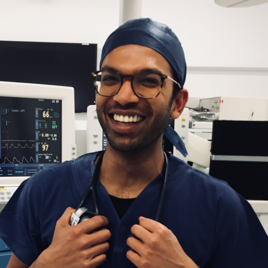 A smiling male healthcare professional in scrubs and a surgical cap, holding a stethoscope in a medical setting with monitors and medical equipment in the background.