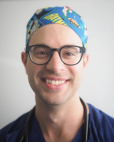 A smiling man wearing glasses and a colorful scrub cap with cartoon characters.